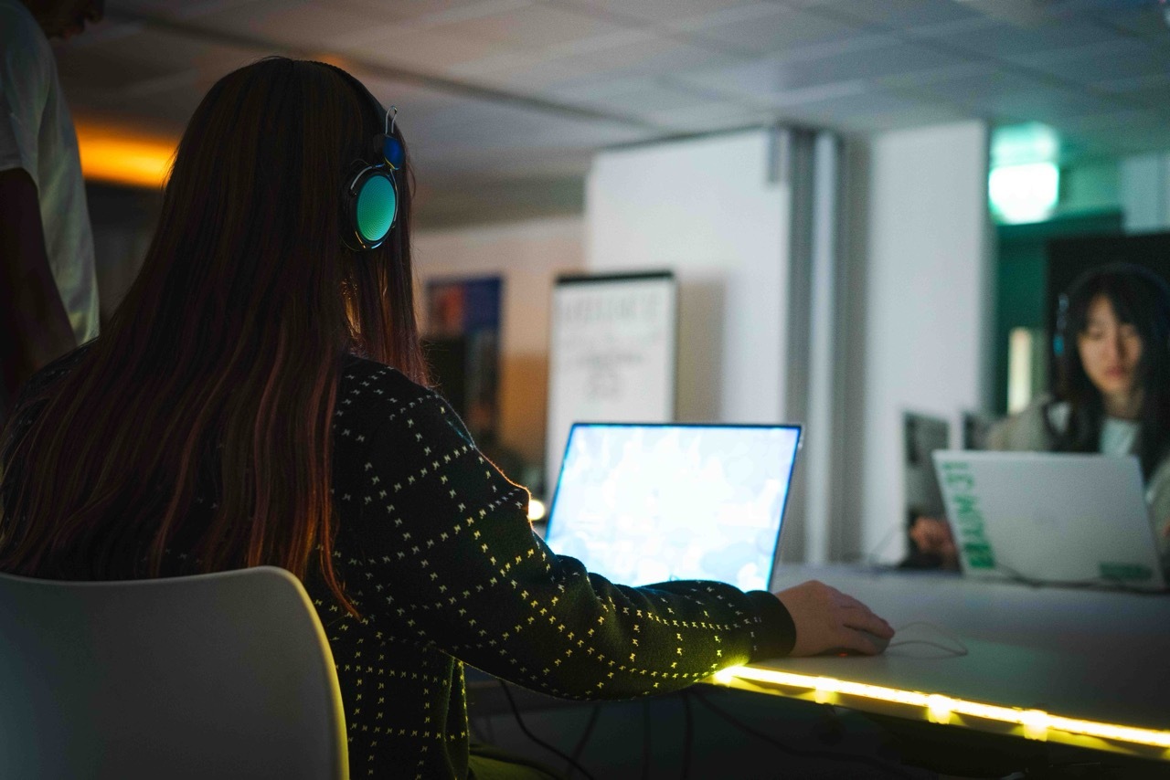 Students interacting and looking at a laptop as part of the exhibition, wearing headphones, yellow LED strip on the table below.