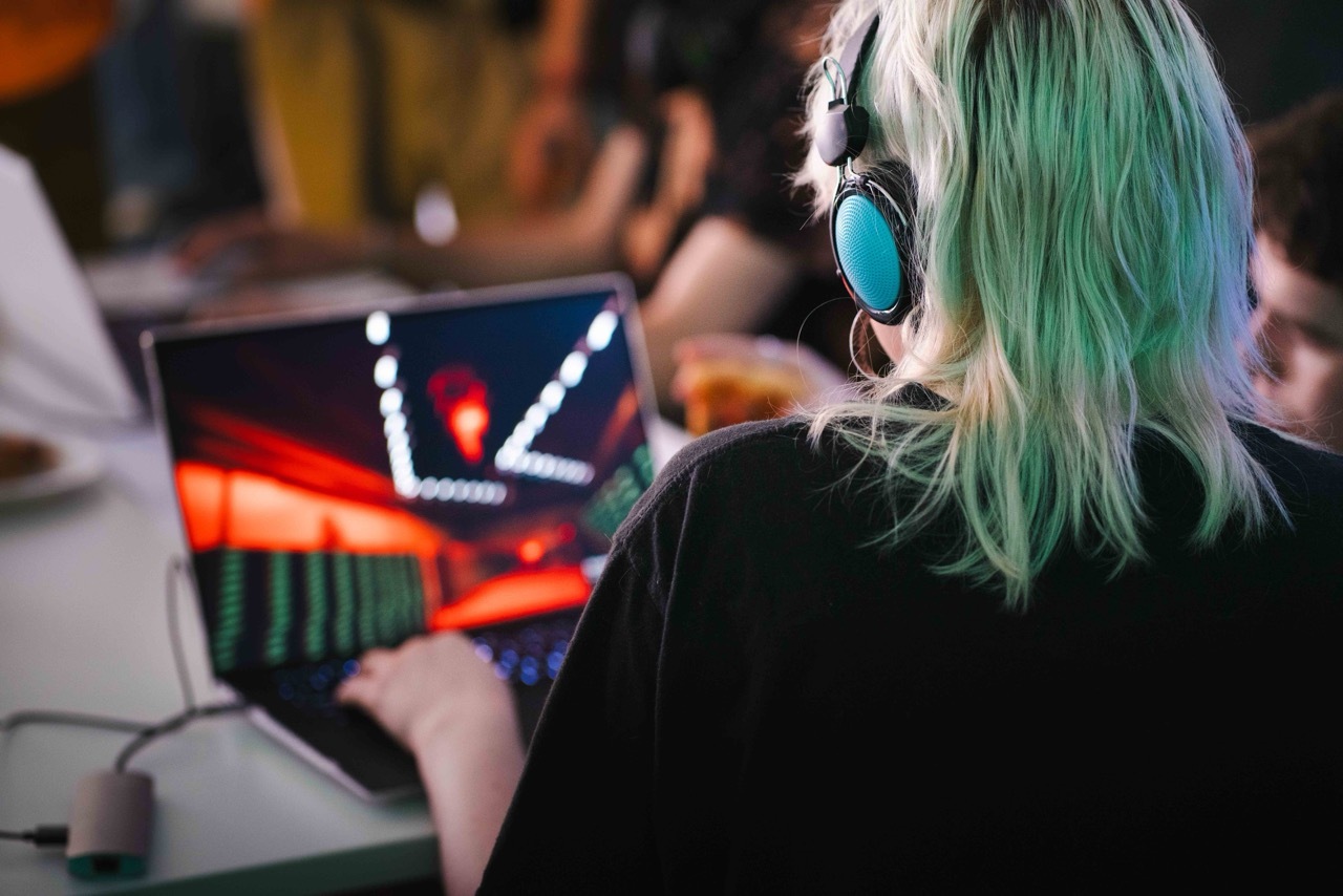 Person with green hair wearing blue headphones playing video game on laptop computer in crowded indoor setting.