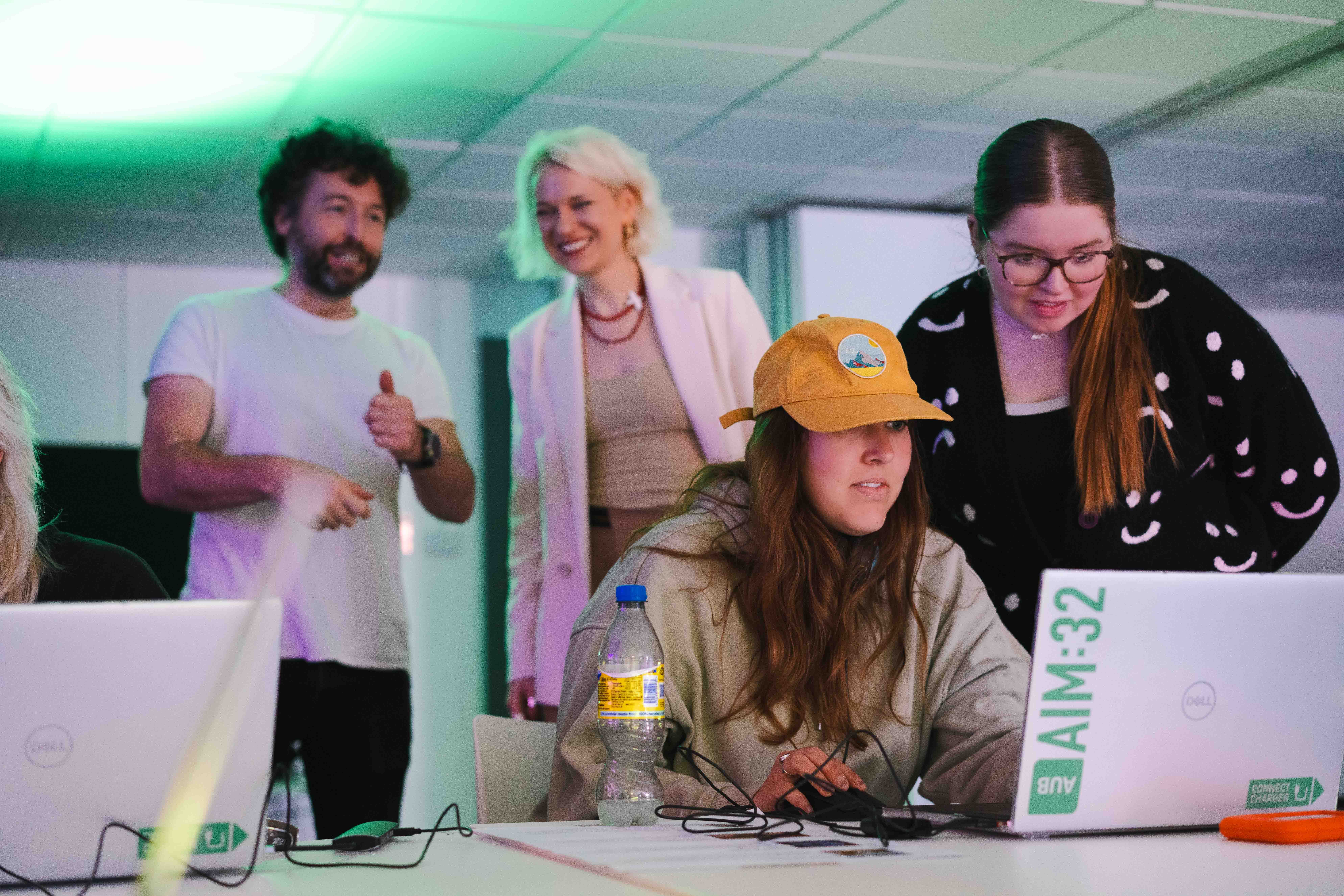 Two Arts University Bournemouth students sitting and playing a videogame on a laptop, with BA (Hons) Games Art and Design staff A. Potemska and J Cutler in the background.
