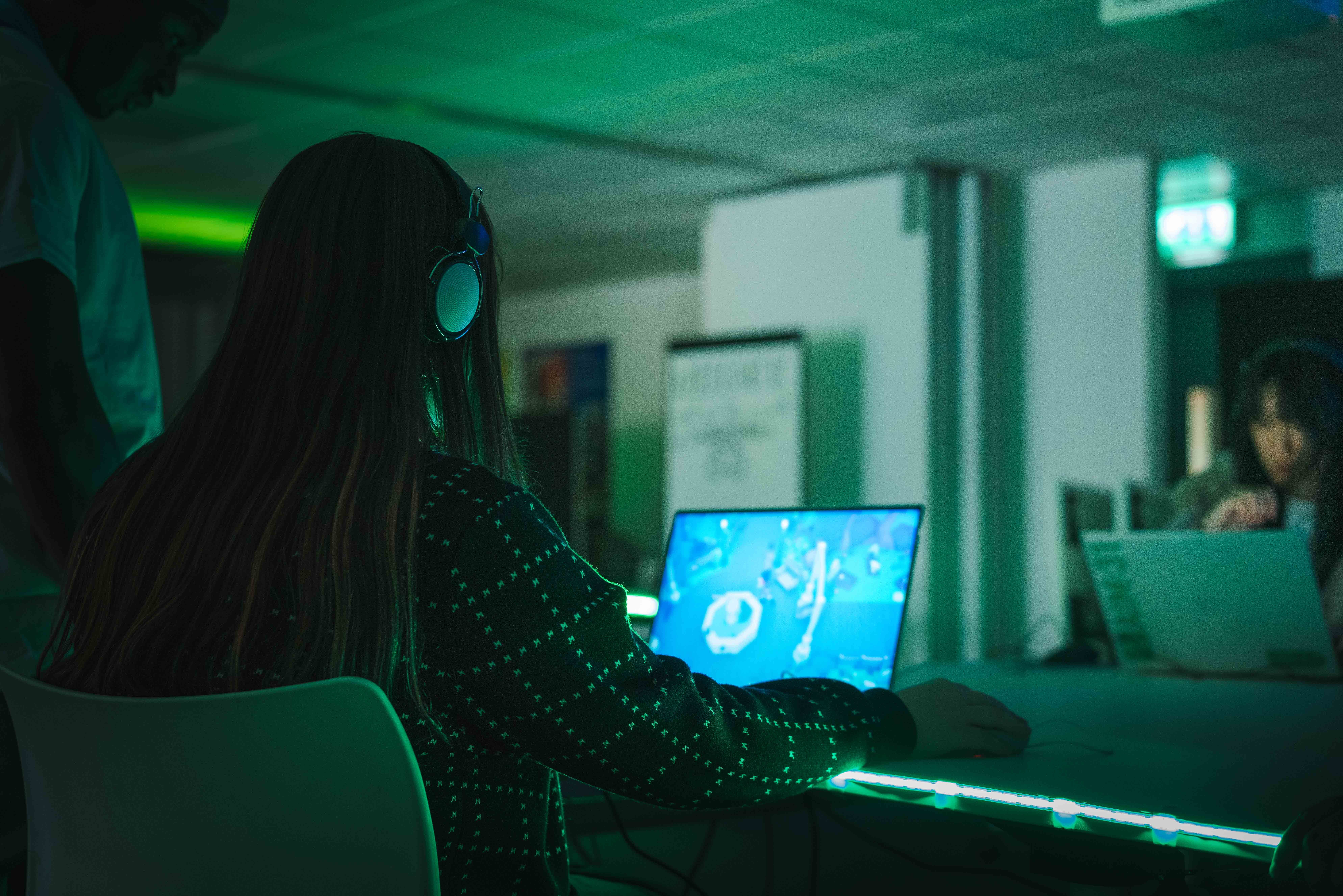 An Arts University Bournemouth student, viewed from the back, sits playing a video game on a laptop at a BA (Hons) Games Art and Design exhibition.