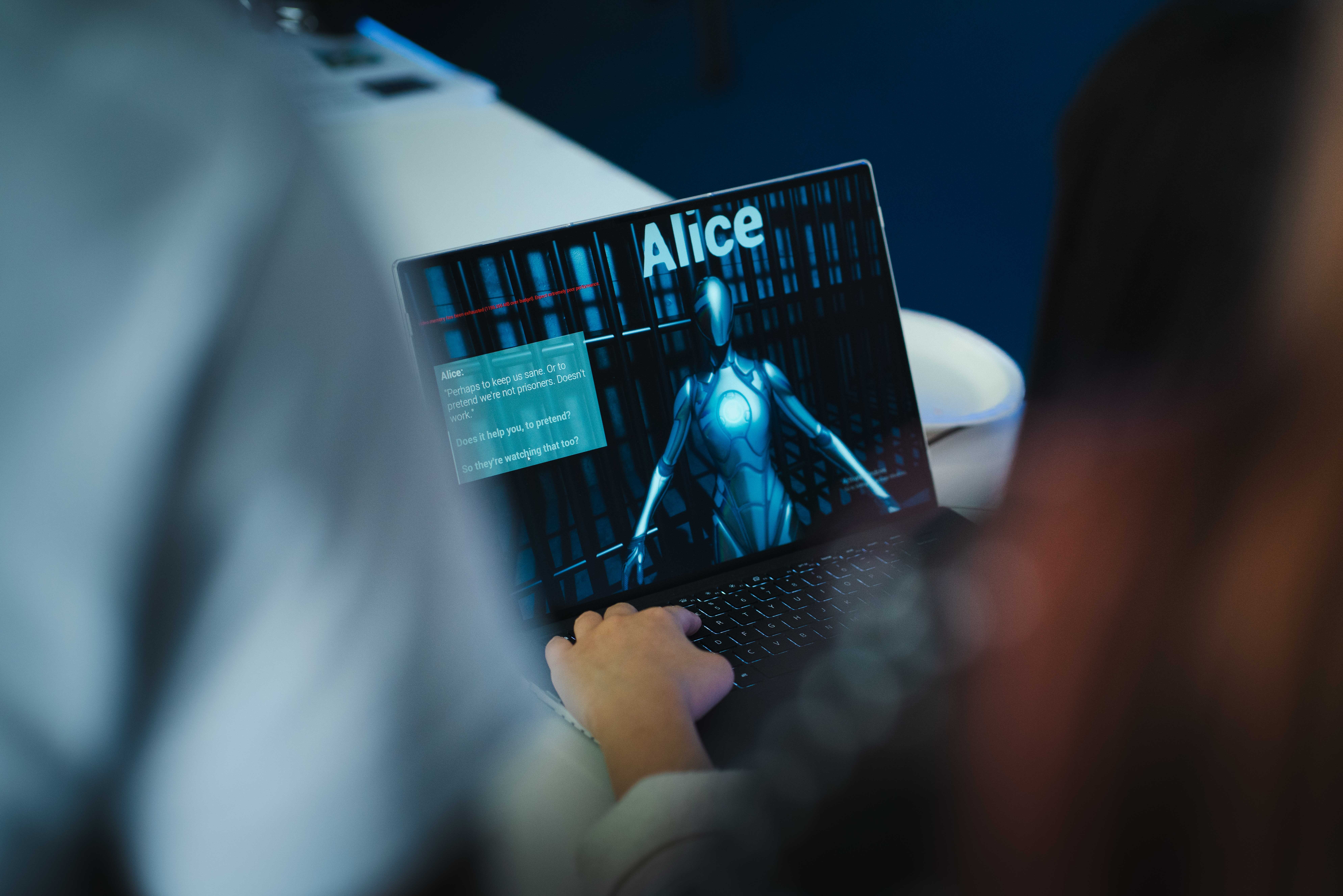 Close-up of a BA (Hons) Games Art and Design student's laptop screen showing a still from a video game – a robot with the name "Alice" above its head, and a dialogue box and text on the left-hand side.