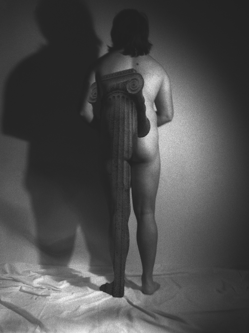 Black and white image of nude figure from behind holding classical column, dramatic lighting creates strong shadows on wall.