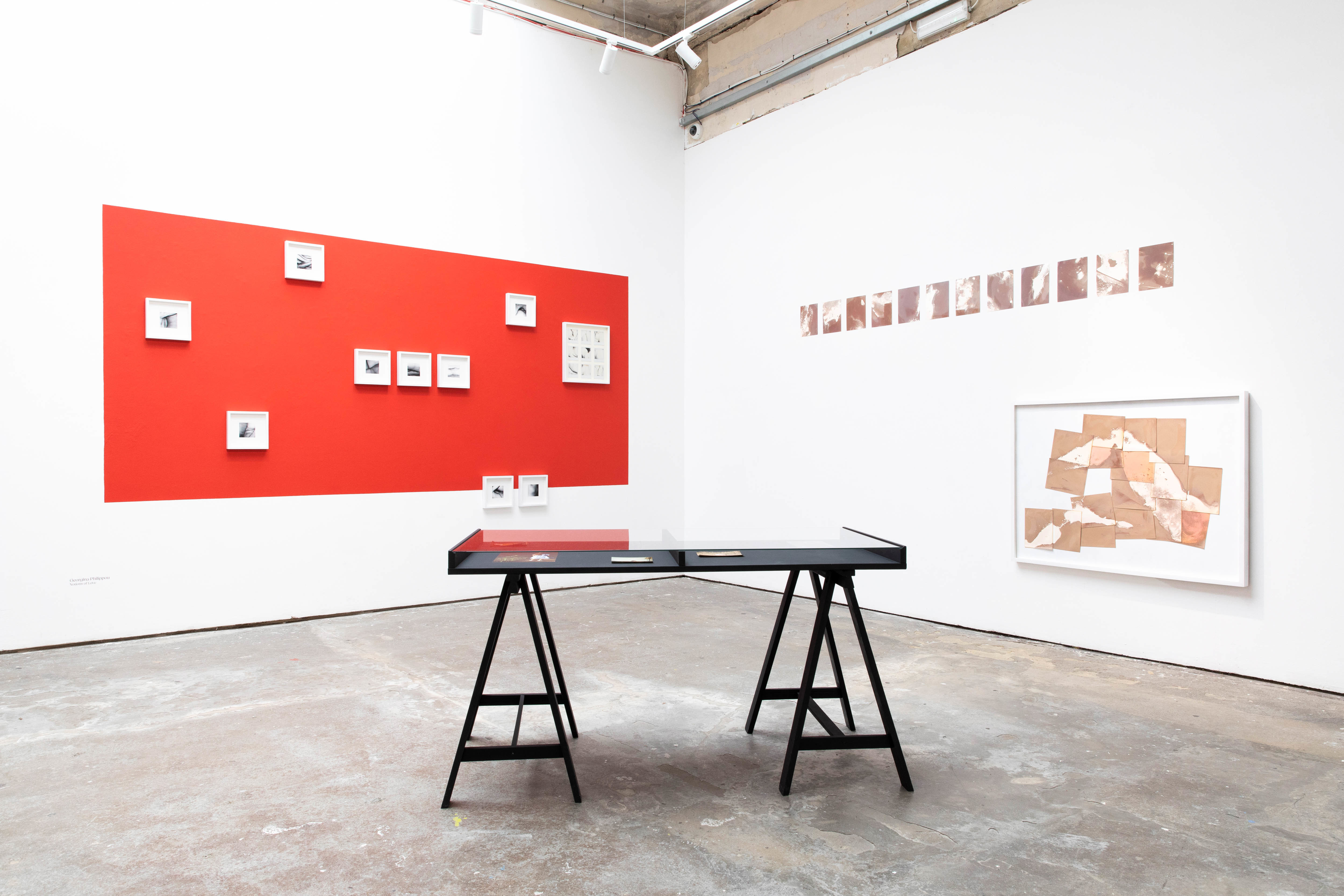 Several miniature artworks in square framed displayed on a wall with a large red rectangle on it. A row of miniature images is displayed on the opposite wall, with a framed abstract artwork hanging underneath. A display case stands in the centre of the space.