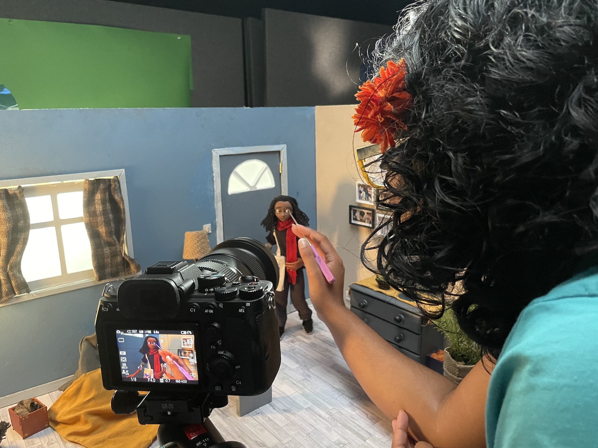 K Roy-Meighoo viewed from the back, working on a stop motion animated character model, in a miniature set of a house's living room. A camera is set up viewing a close-up of the animated character on the monitor.