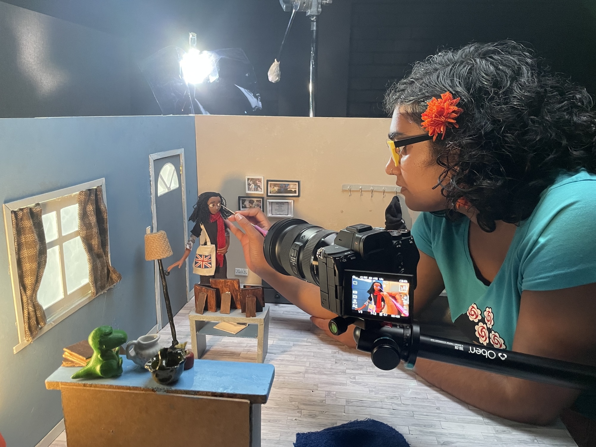 K Roy-Meighoo working on a stop motion animated figure, in a miniature set of a house's living room. A camera is set up viewing a close-up of the animated character on the monitor.