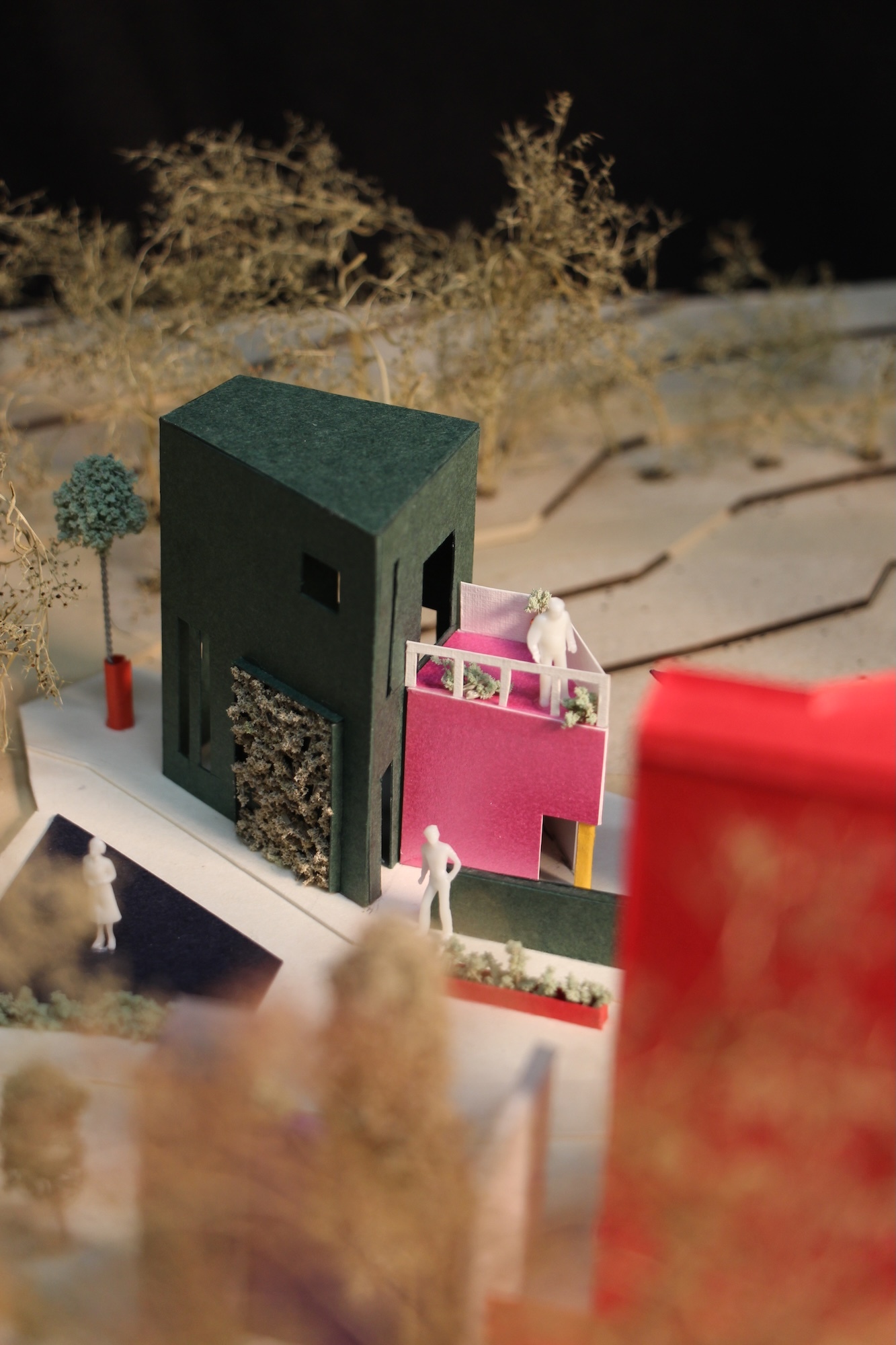 Miniature architectural model with dark green and pink geometric buildings on white platform, small white figures, dried vegetation behind.