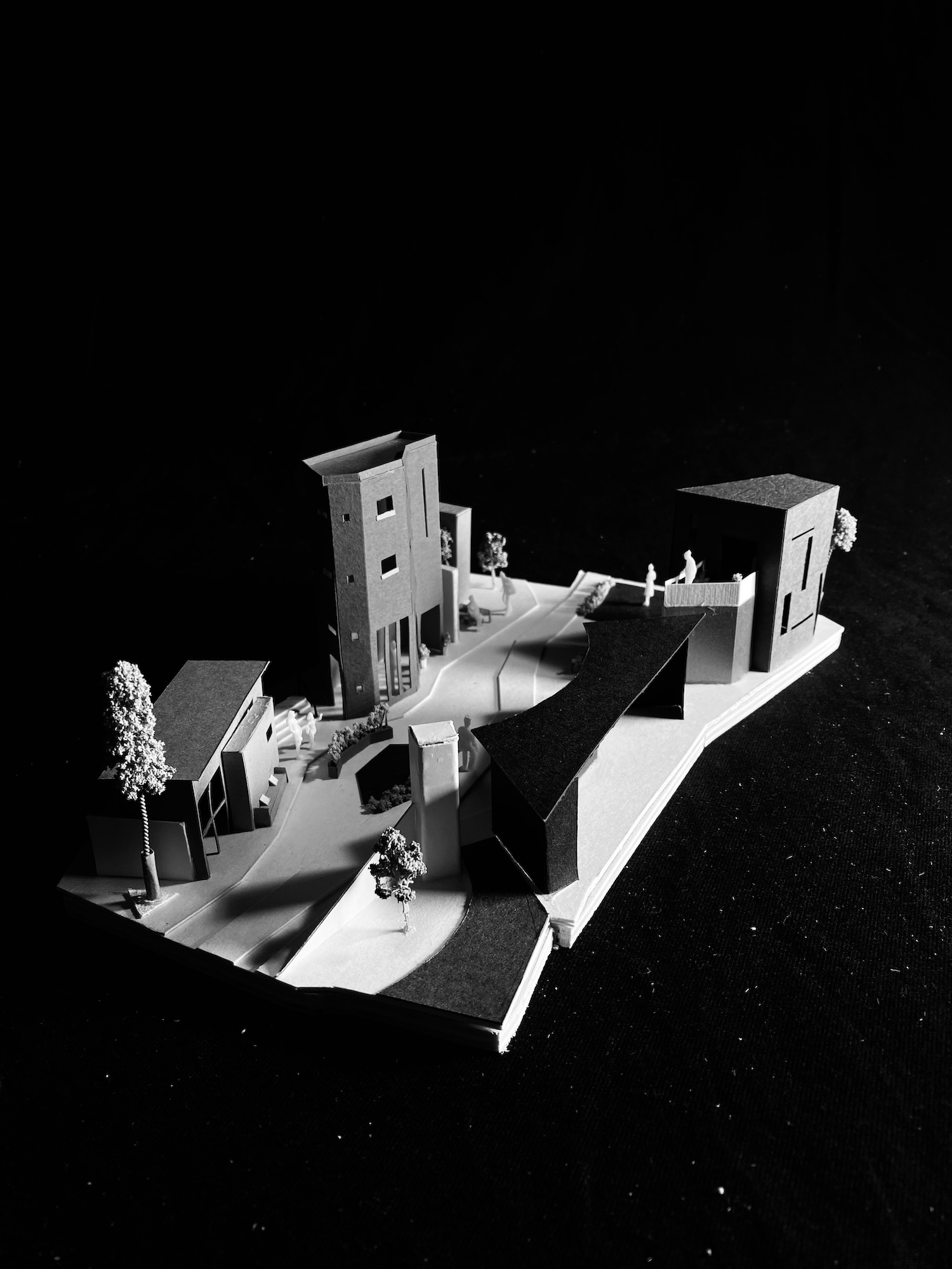 White architectural model with angular buildings, curved pathways, and small figures on black background.