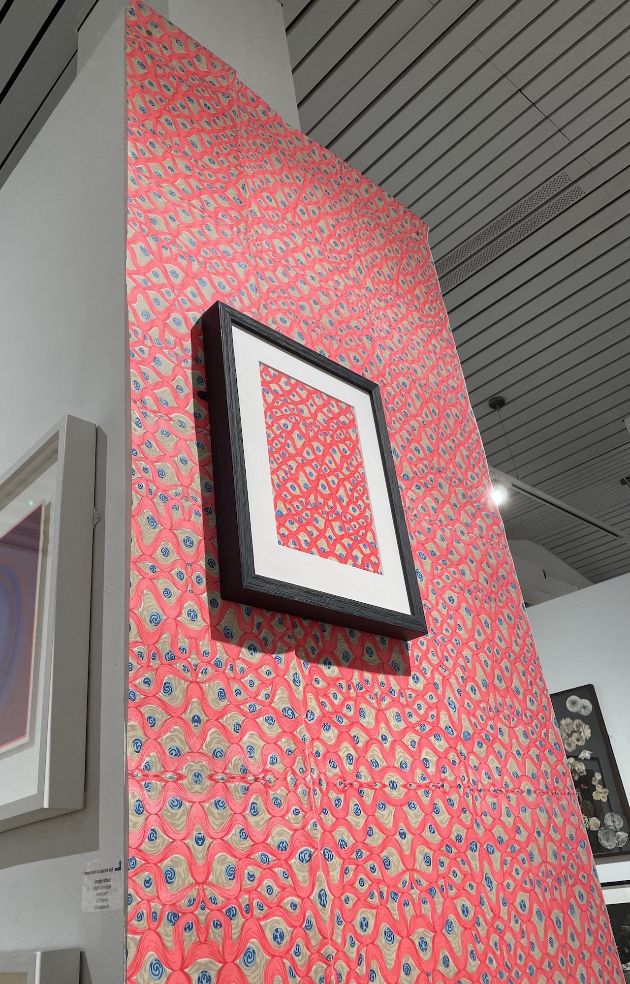 A wall of the Bankside Gallery lined with colourful pink and patterned wallpaper. A sample of the same wallpaper is displayed in a frame on top of it.