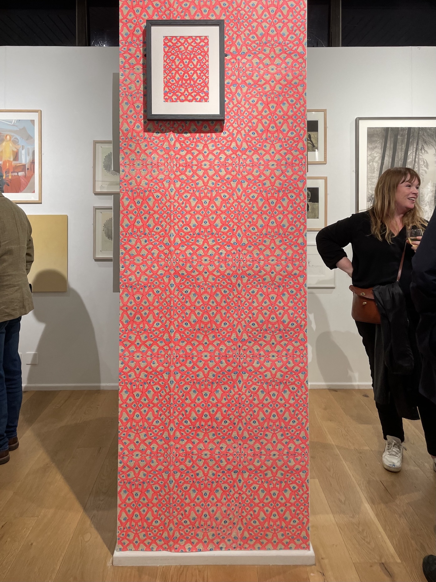 A wall of the Bankside Gallery lined with colourful pink and patterned wallpaper. A sample of the same wallpaper is displayed in a frame on top of it. Gallery visitors are seen standing either side of the wall.