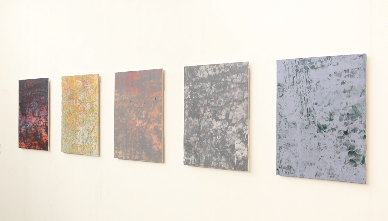 Five abstract paintings hung on white wall in gallery setting, featuring muted colours ranging from burgundy to grey to pale blue.
