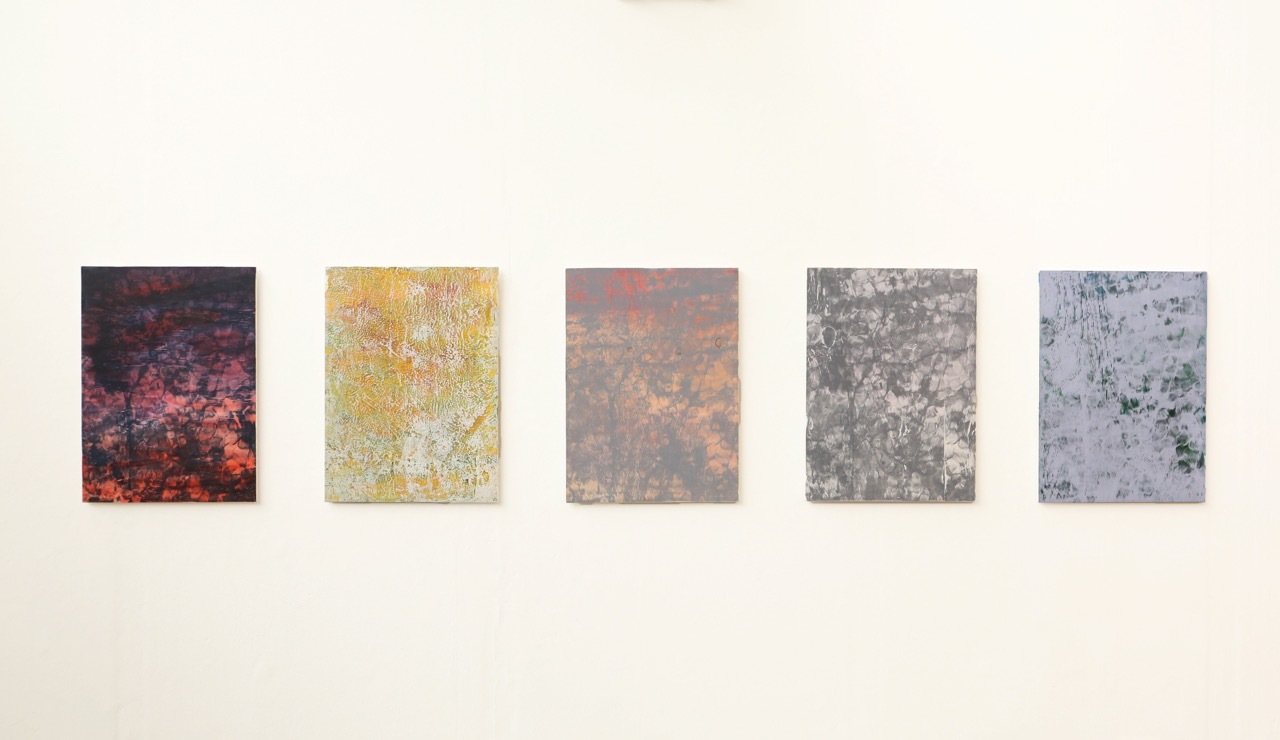 Five rectangular artworks mounted on white wall in horizontal row. Left to right: dark purple-red, bright yellow, brown-orange, grey-green, pale blue-grey. All have textured, mottled surfaces.
