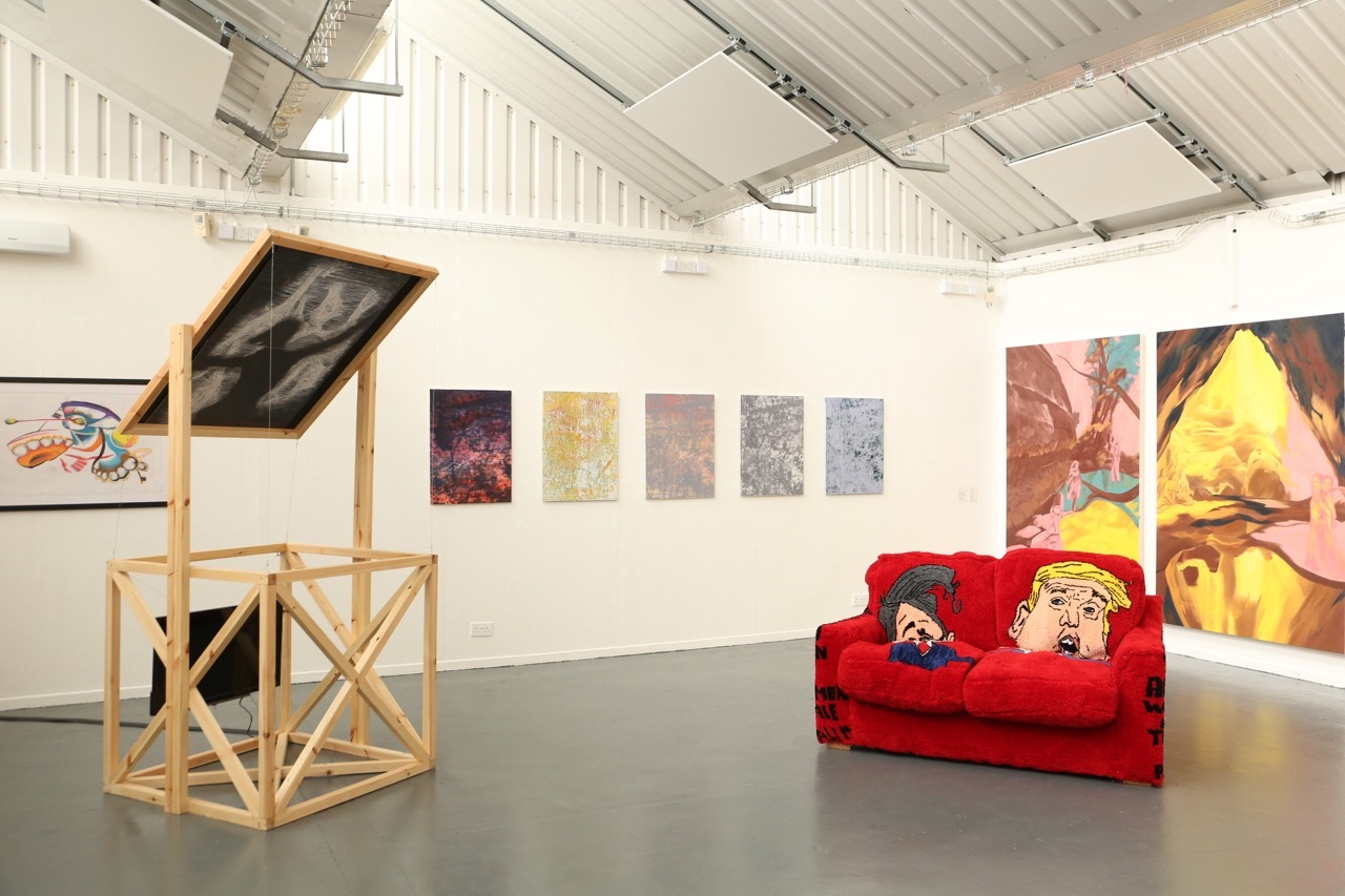 White-walled gallery with exposed ceiling beams, wooden sculpture structure, red sofa, and various artworks including colourful paintings.