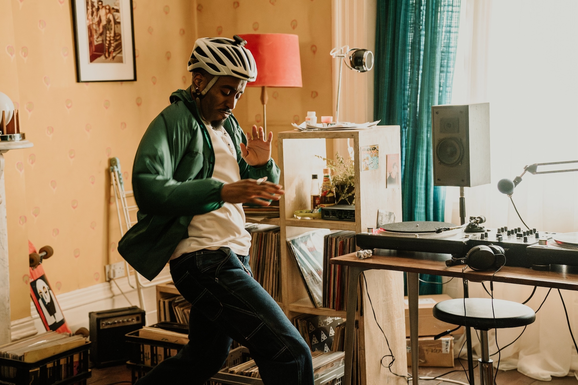 A man posing mid-dance while wearing a bike helmet, next to a desk and DJ deck.