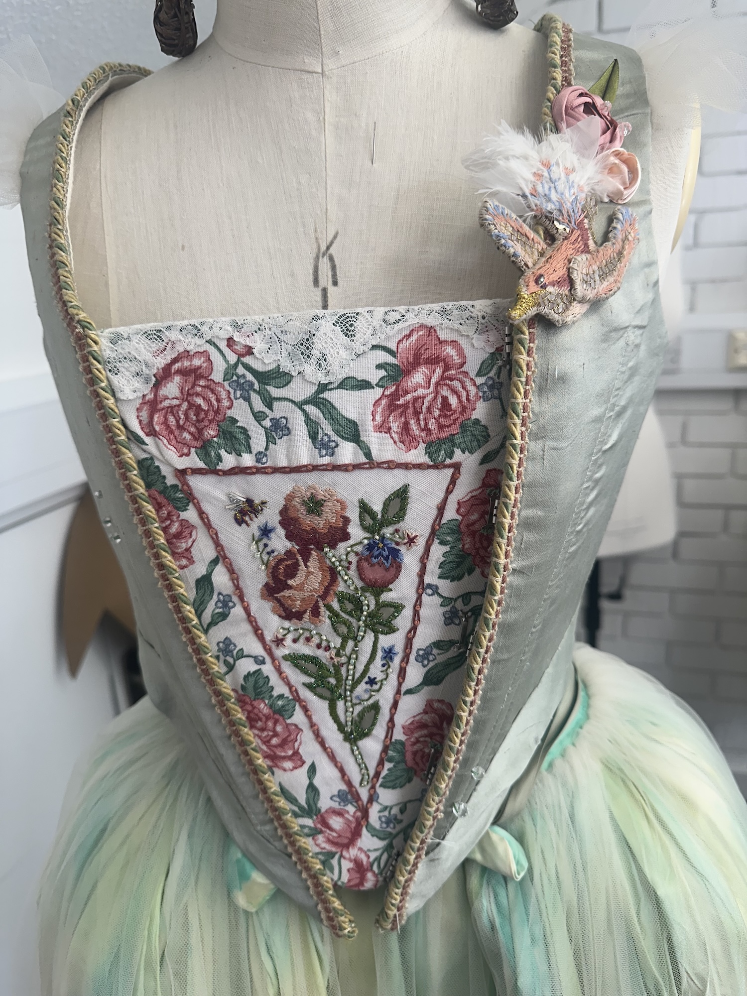 A dressmaker's mannequin wearing a pastel costume with a floral embroidered bodice, gold braid trim and a layered tulle skirt, with a decorative flower on the shoulder.