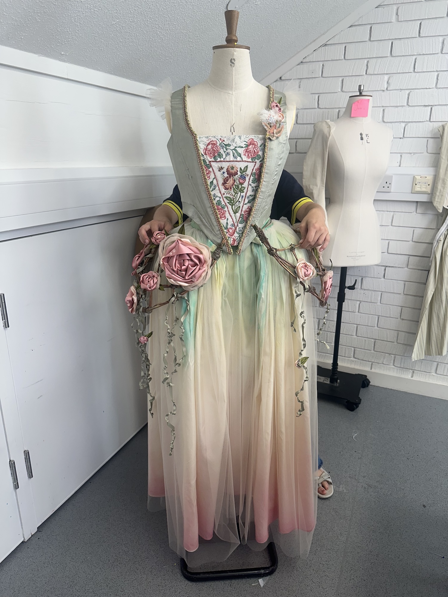 A dress form displays a pastel tulle gown with floral embroidery and rose decorations, set in a sewing studio with white cabinets and another mannequin in the background.