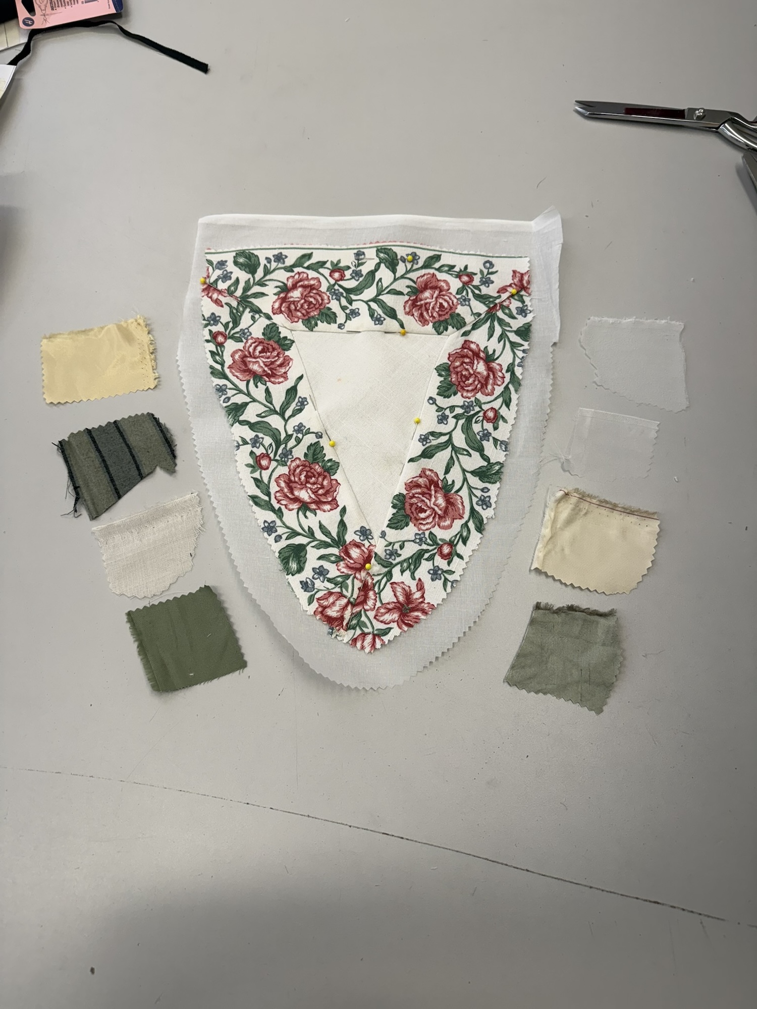 A floral fabric garment piece with red roses and green leaves is laid on a grey table, surrounded by small fabric swatches and scissors.