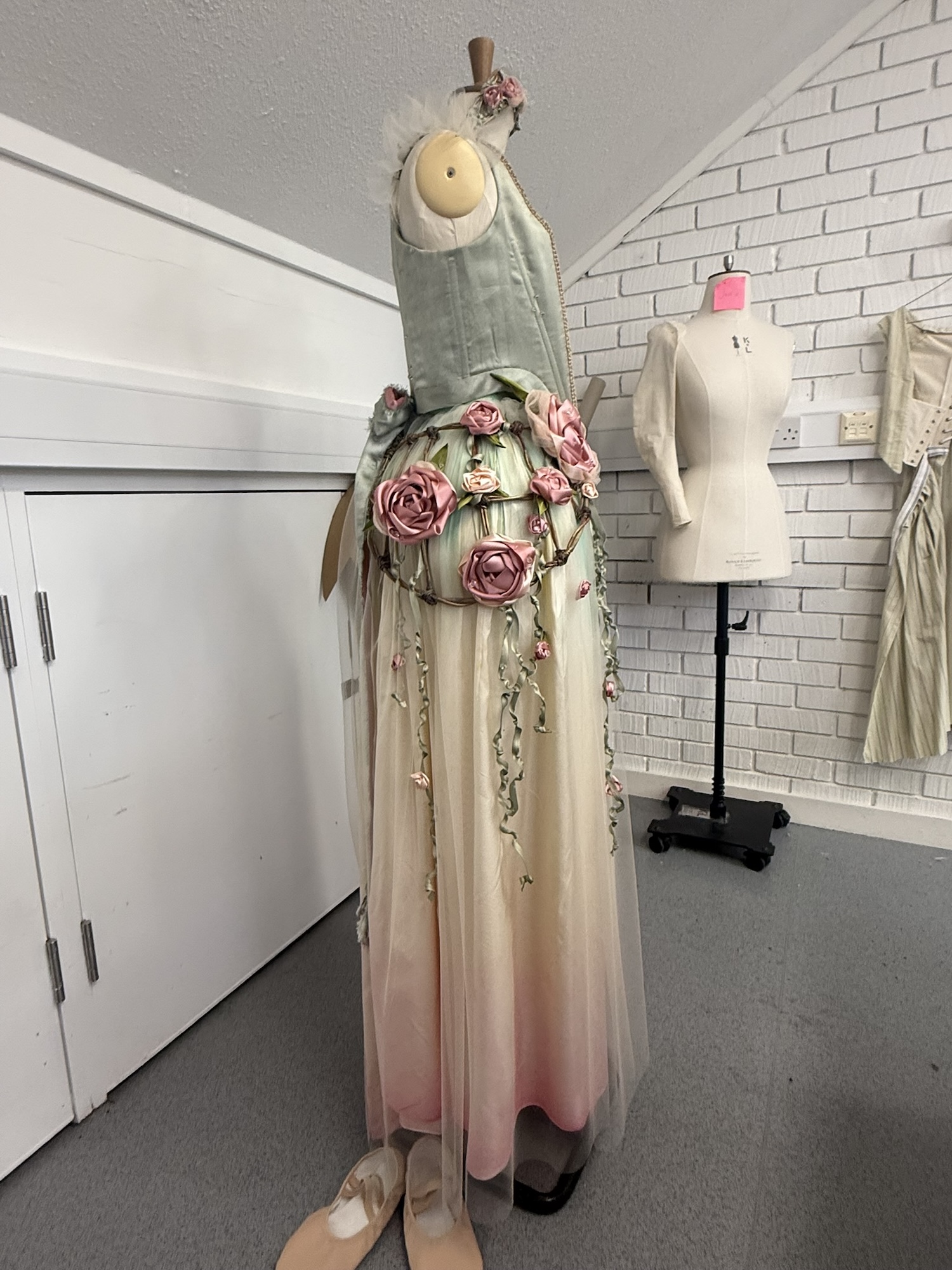 A dress form displays a long pastel gown with floral decorations and trailing ribbons, standing in a sewing room beside a second mannequin and white cabinets.