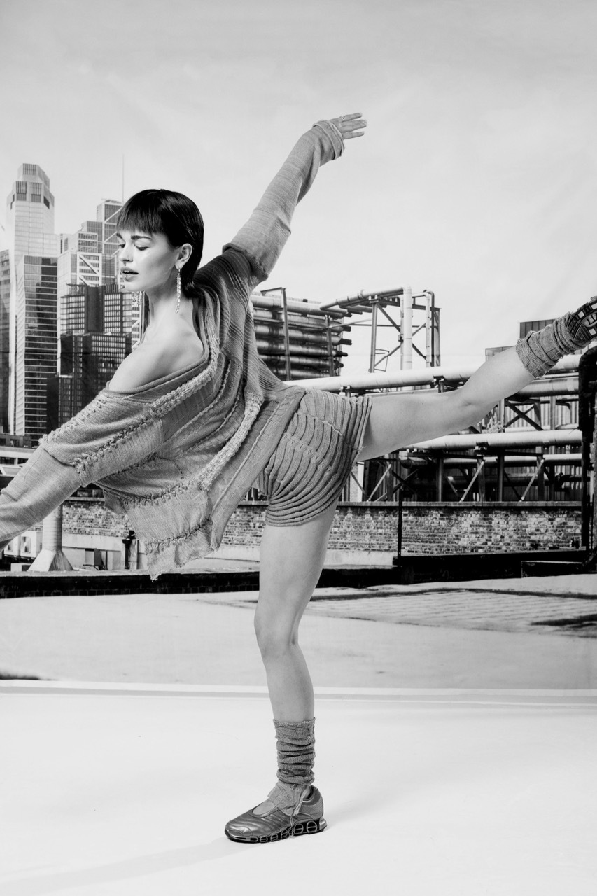 Black and white image of woman in fringed dress performing high kick dance move on rooftop with city skyline behind.