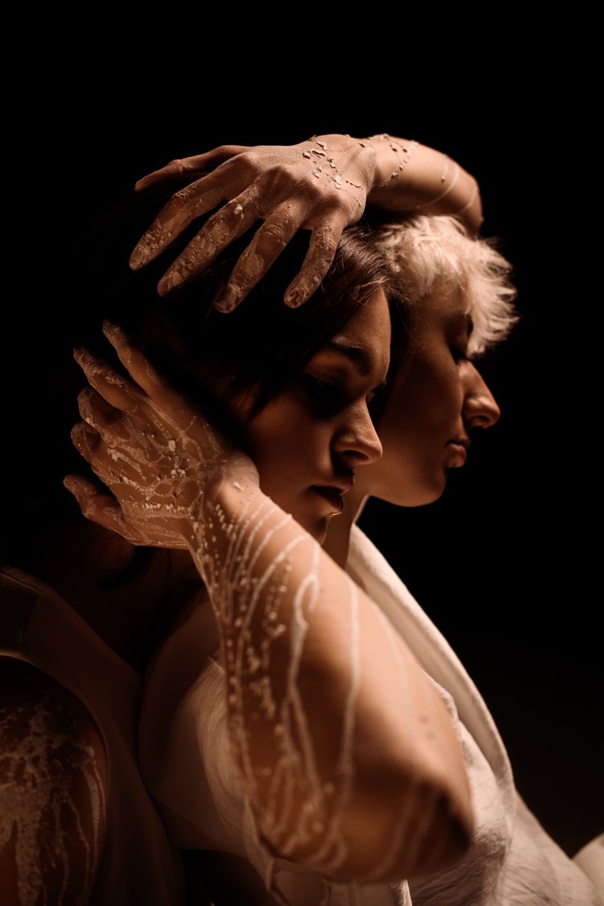 Two people in profile with dirt droplets covering their skin and hair, hands positioned near their faces against black background.