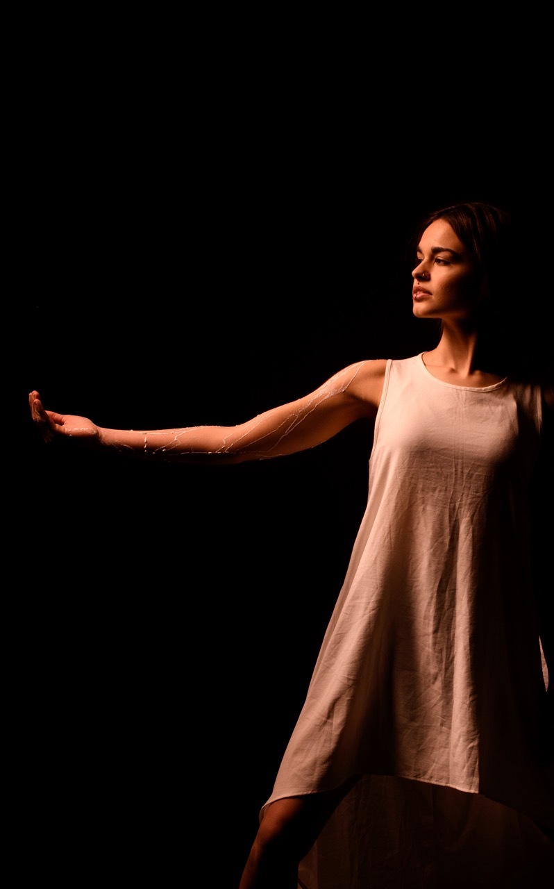 Woman in white sleeveless dress with arms outstretched, lit by warm orange stage lighting against black background.