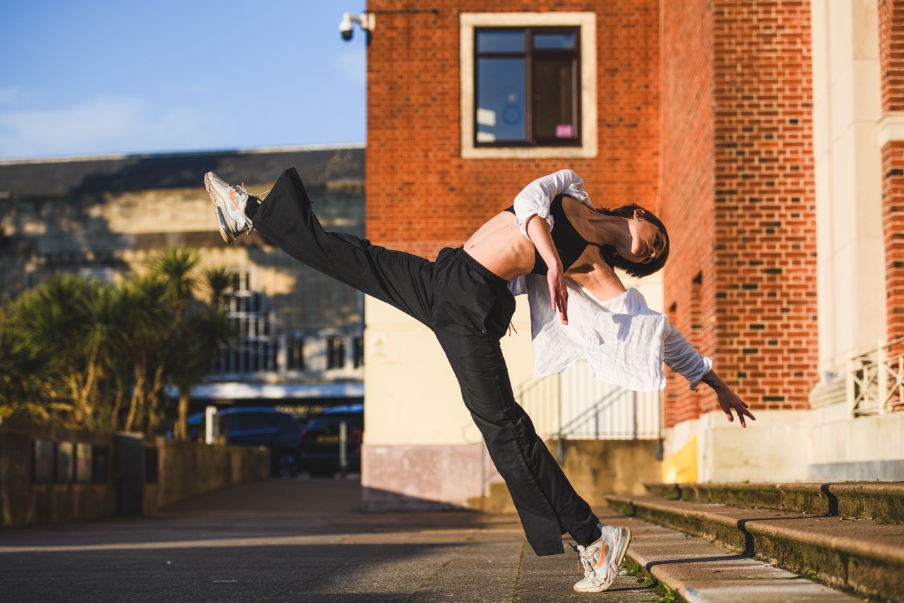 Woman in black leggings and white top performing dynamic dance move with extended leg against red brick building backdrop.
