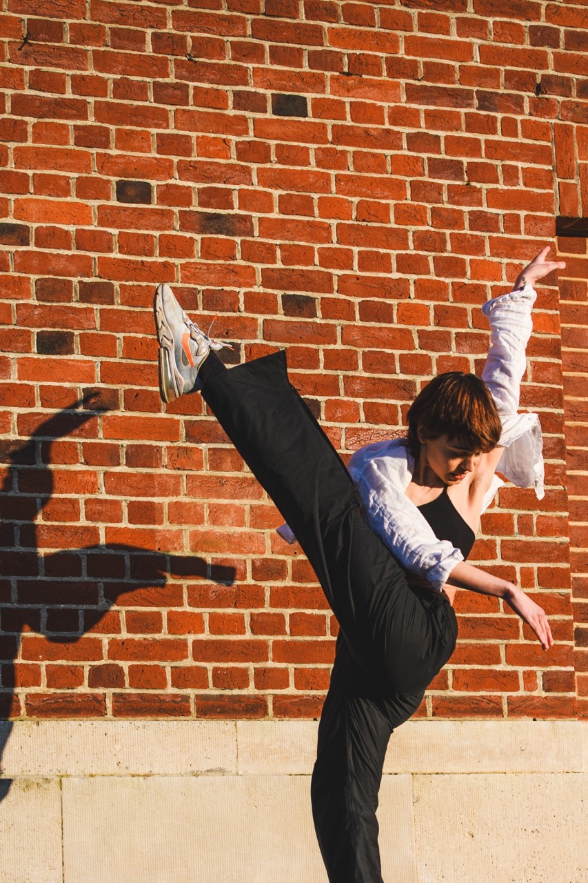 Person in black trousers and white shirt performing high kick against red brick wall with white mortar joints.