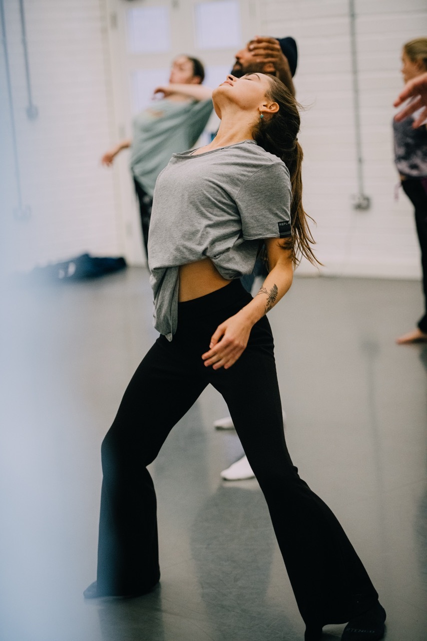 Woman in grey top and black trousers dancing with head tilted back, ponytail swinging. Other dancers visible in background of studio.