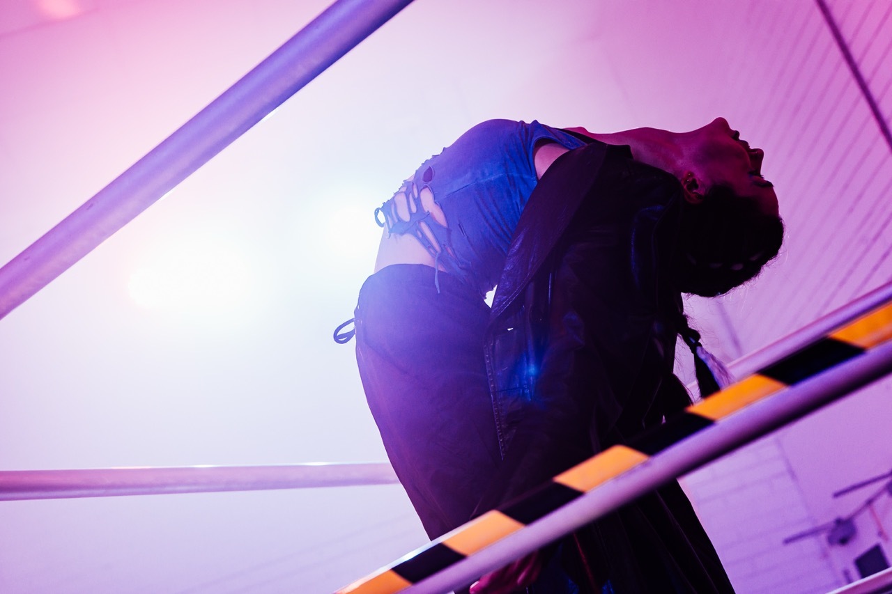 Person with curly hair silhouetted against bright white light, surrounded by purple- and pink-illuminated barriers.