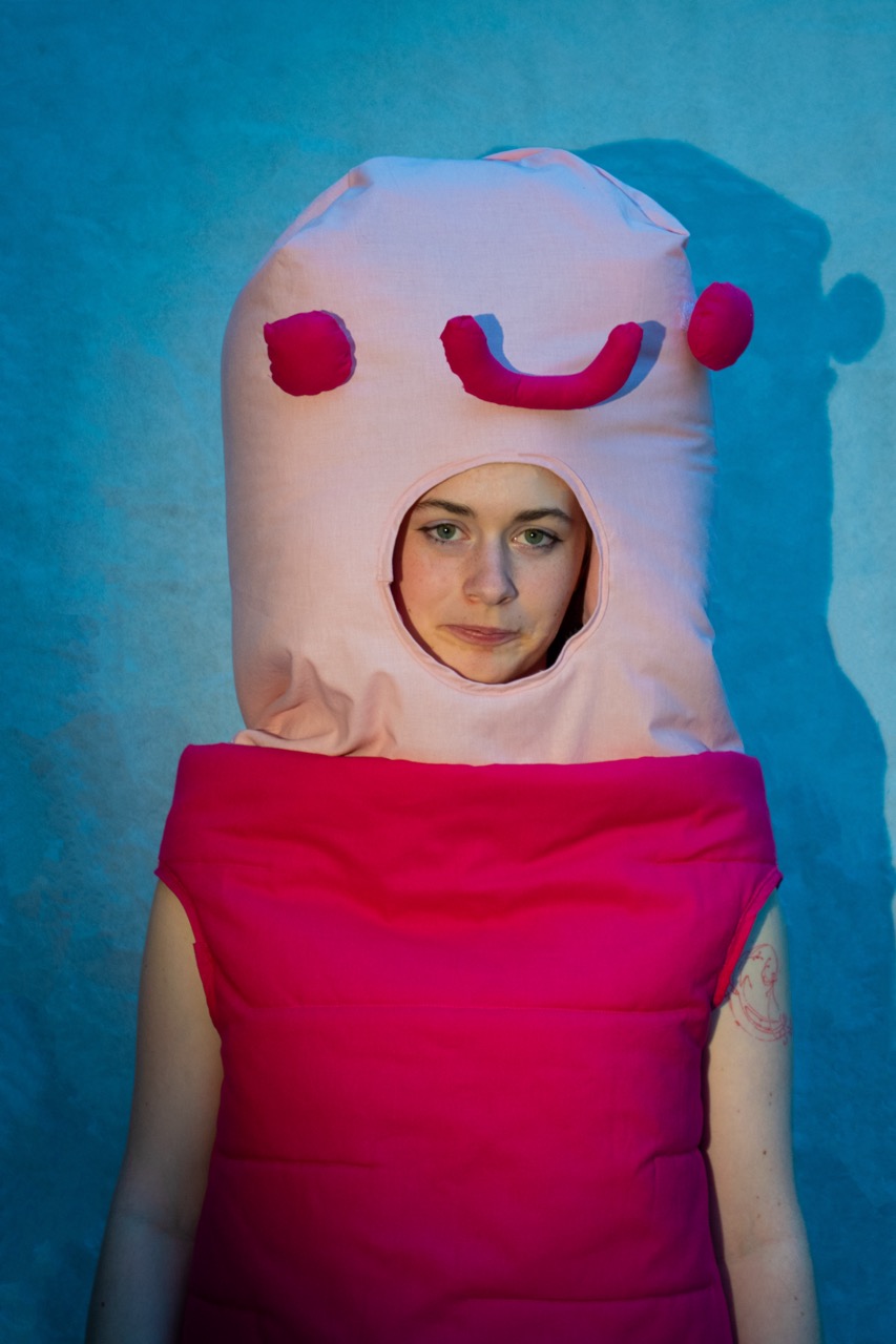 A person wearing a pink costume with a large padded headpiece stands against a blue background, with a face opening in the centre and a shadow on the wall.