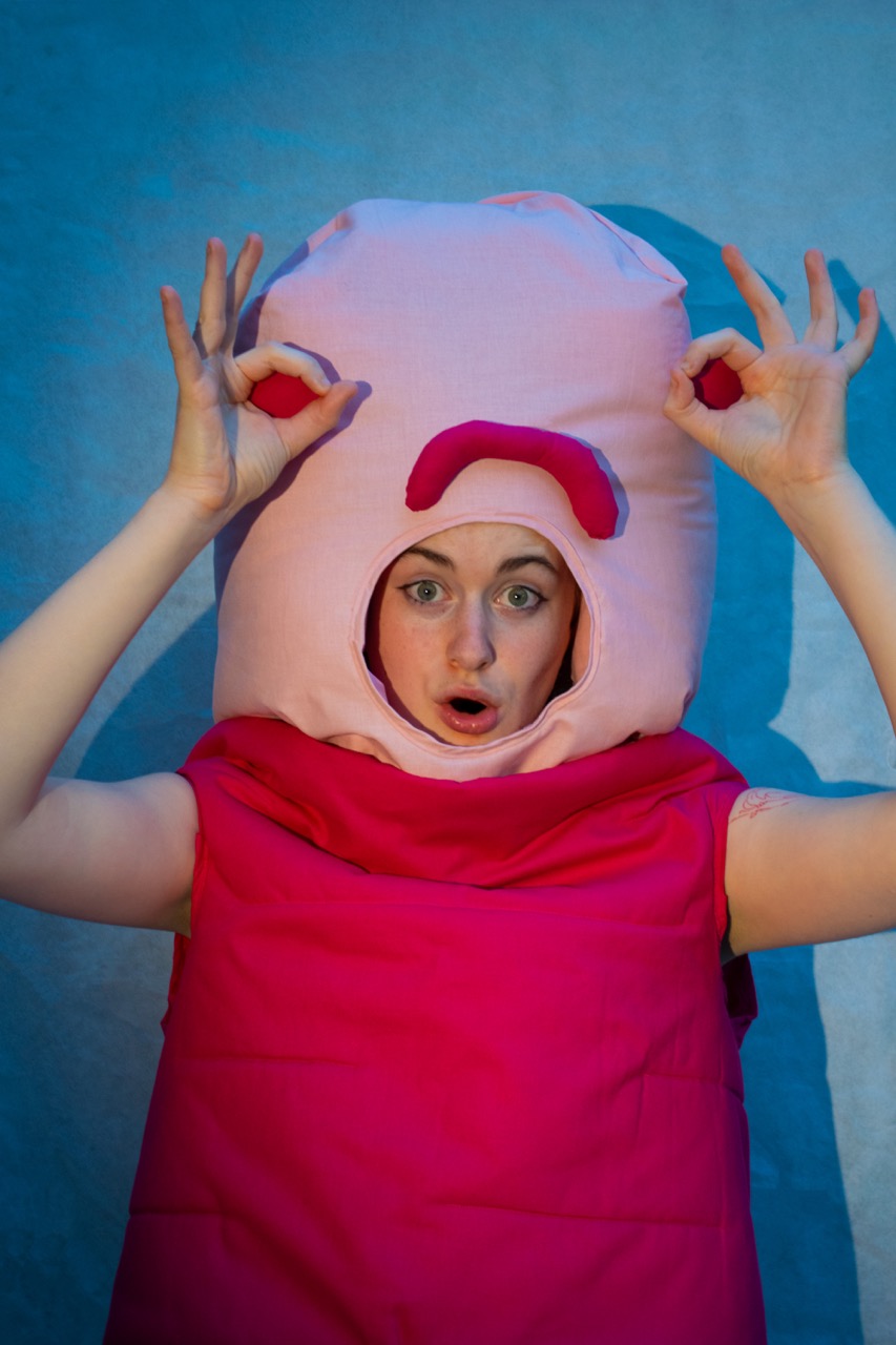 A woman dressed as a pink costume character poses against a blue background, holding both hands near her head.