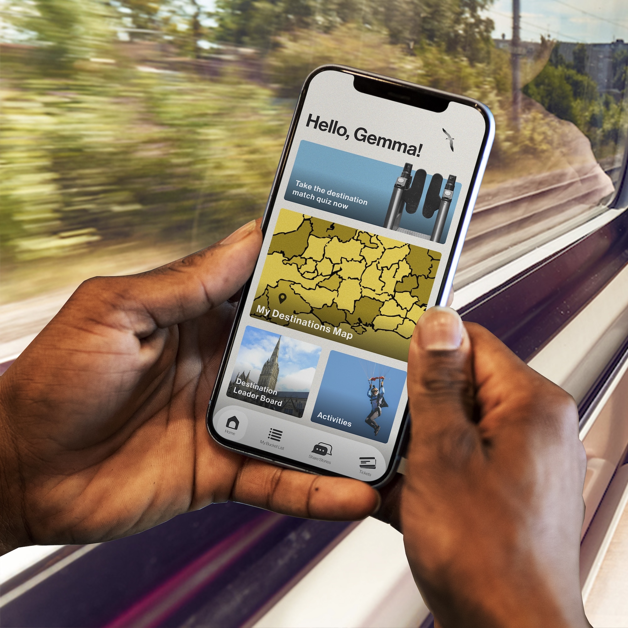 Hands holding smartphone displaying travel app with "Hello, Gemma!" greeting, yellow map, and blue destination images, viewed from moving train.