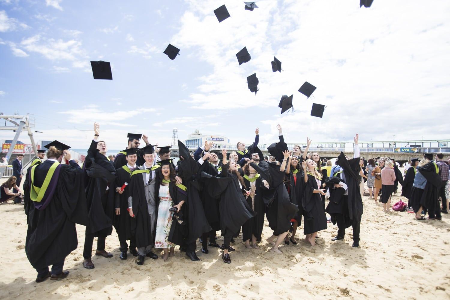 A large group of students standing on a beach in graduation dress throw their mortar board hats in the air