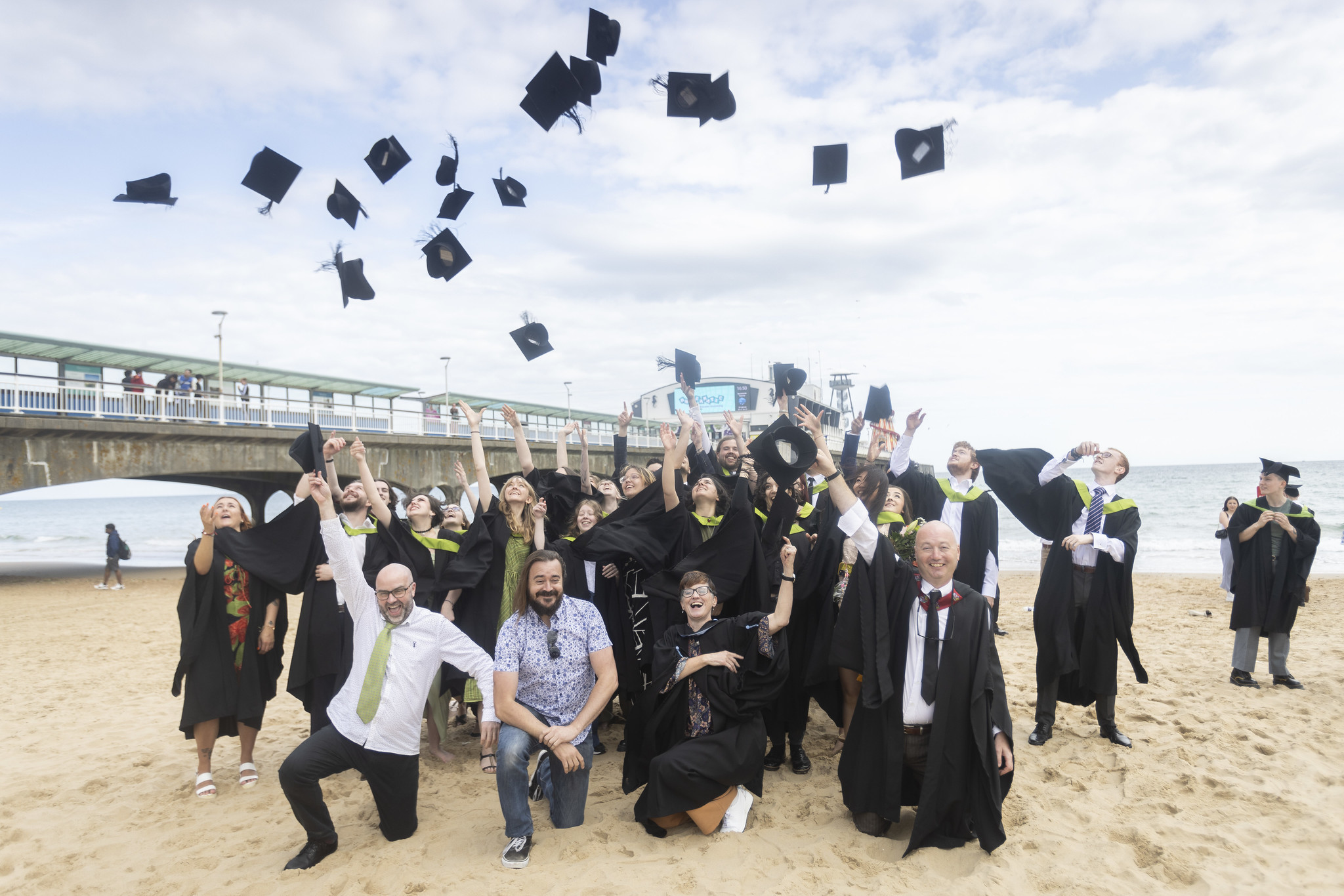 Large group of AUB graduates and lecturers gathers on Bournemouth Beach and throws their mortarboard hats into the air.