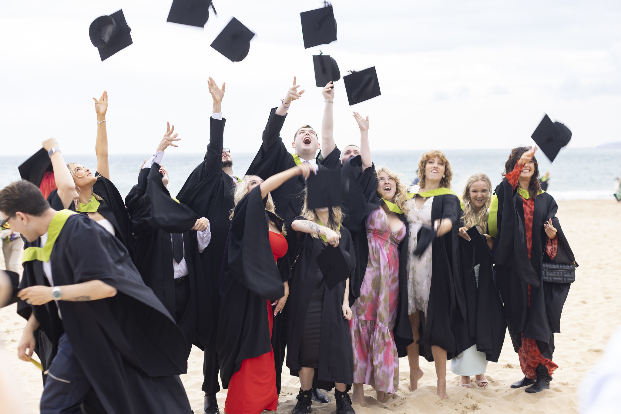 A group of graduates from AUB throw their mortar board hats into the air while standing on Bournemouth Beach