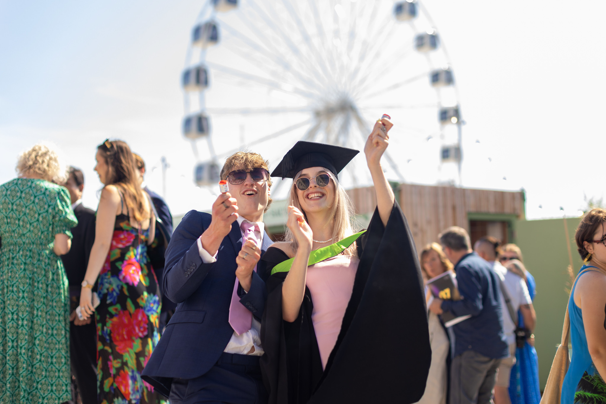 Graduates popping confetti outside, Bournemouth ferris wheel blurred in the background.