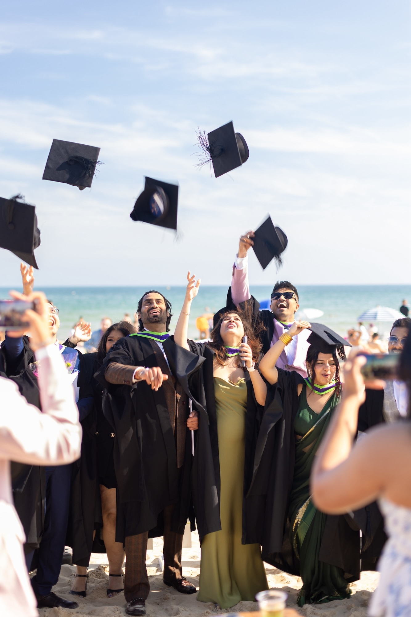 Graduates from Arts University Bournemouth in graduation gowns standing on Bournemouth Beach and throwing their mortar boards in the air.