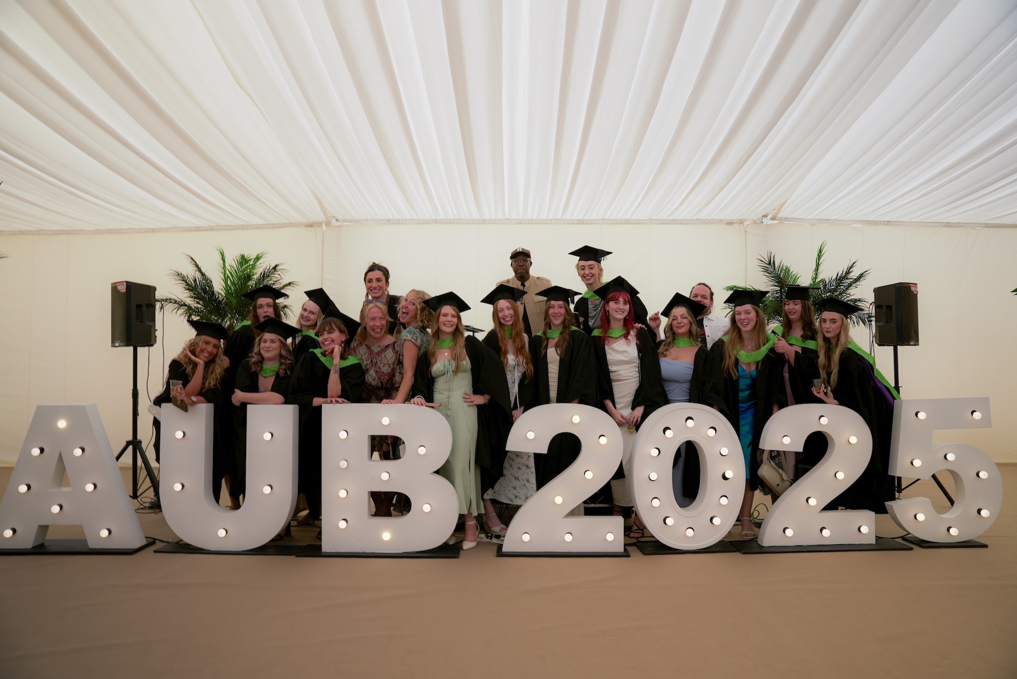 Graduates in caps and gowns pose behind large white "HUB 2022" letters with LED lights in a marquee tent.