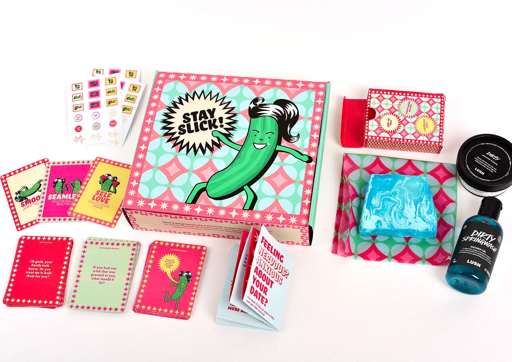 Colourful subscription box contents with pink geometric packaging, cartoon pickle character, beauty products, and assorted cards spread on white surface.