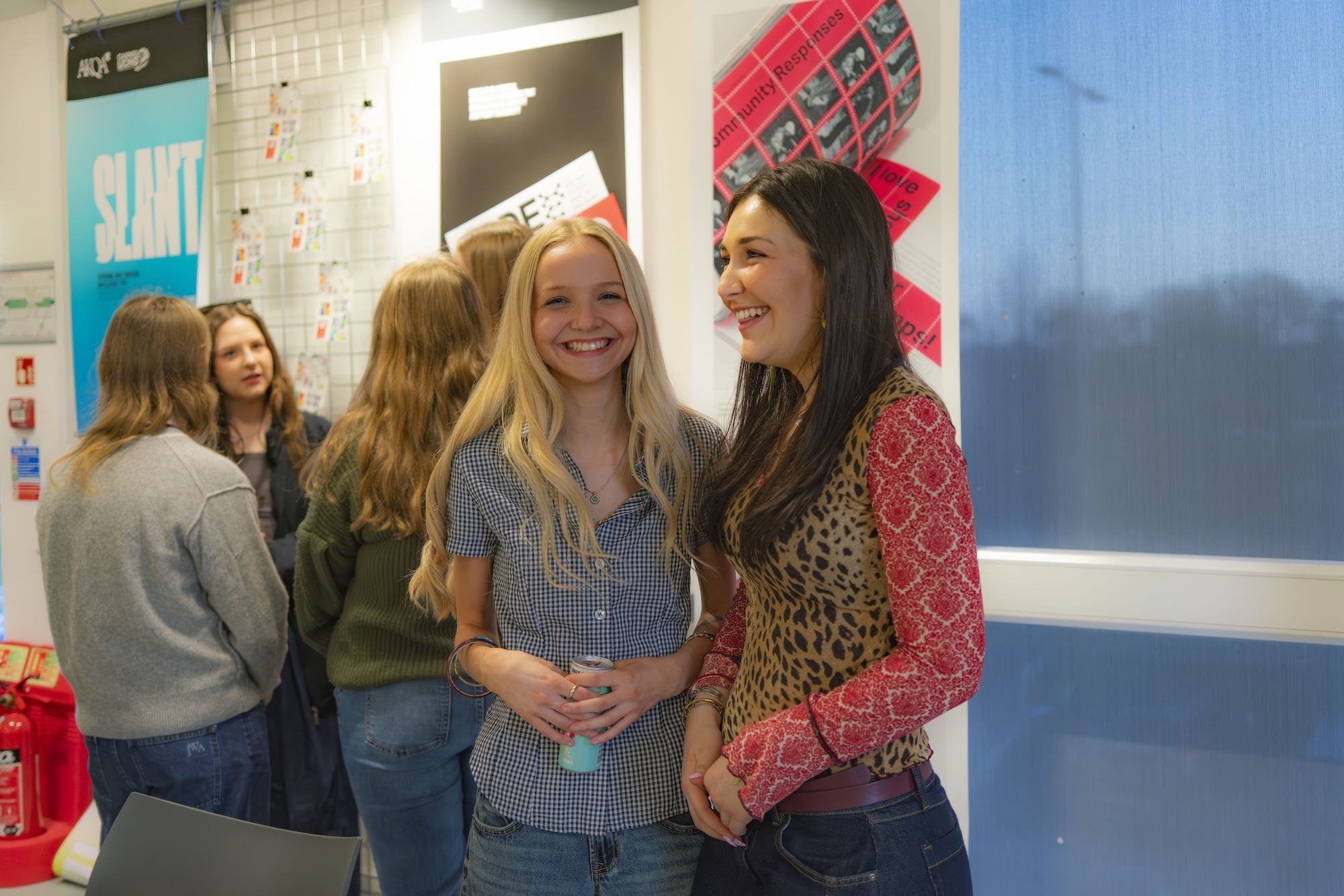 Group of BA (Hons) Graphic Design students socialising in bright classroom with white walls, blue accents, and red decorative elements on ceiling.