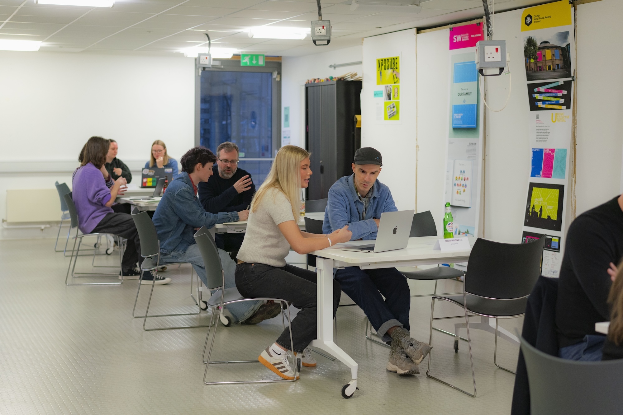 Office meeting room with people seated at white tables with laptops. Colourful post-it notes on white walls, blue doors visible.