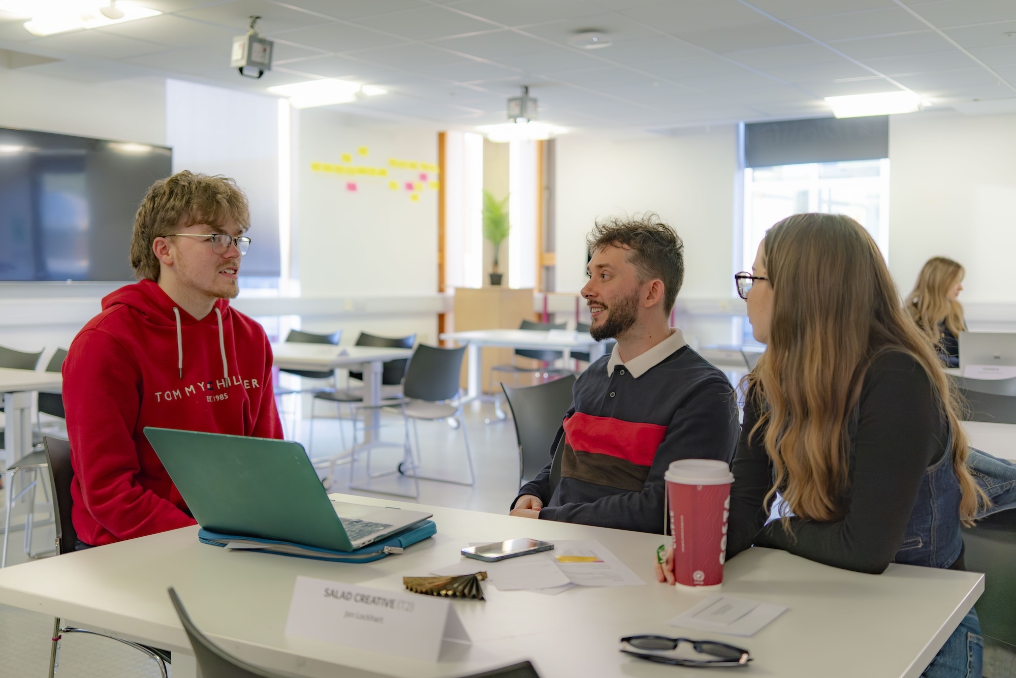 A BA (Hons) Graphic Design students sits with two representatives from Salad Creative at white table in modern classroom with laptops, red cups, and colourful wall displays visible.