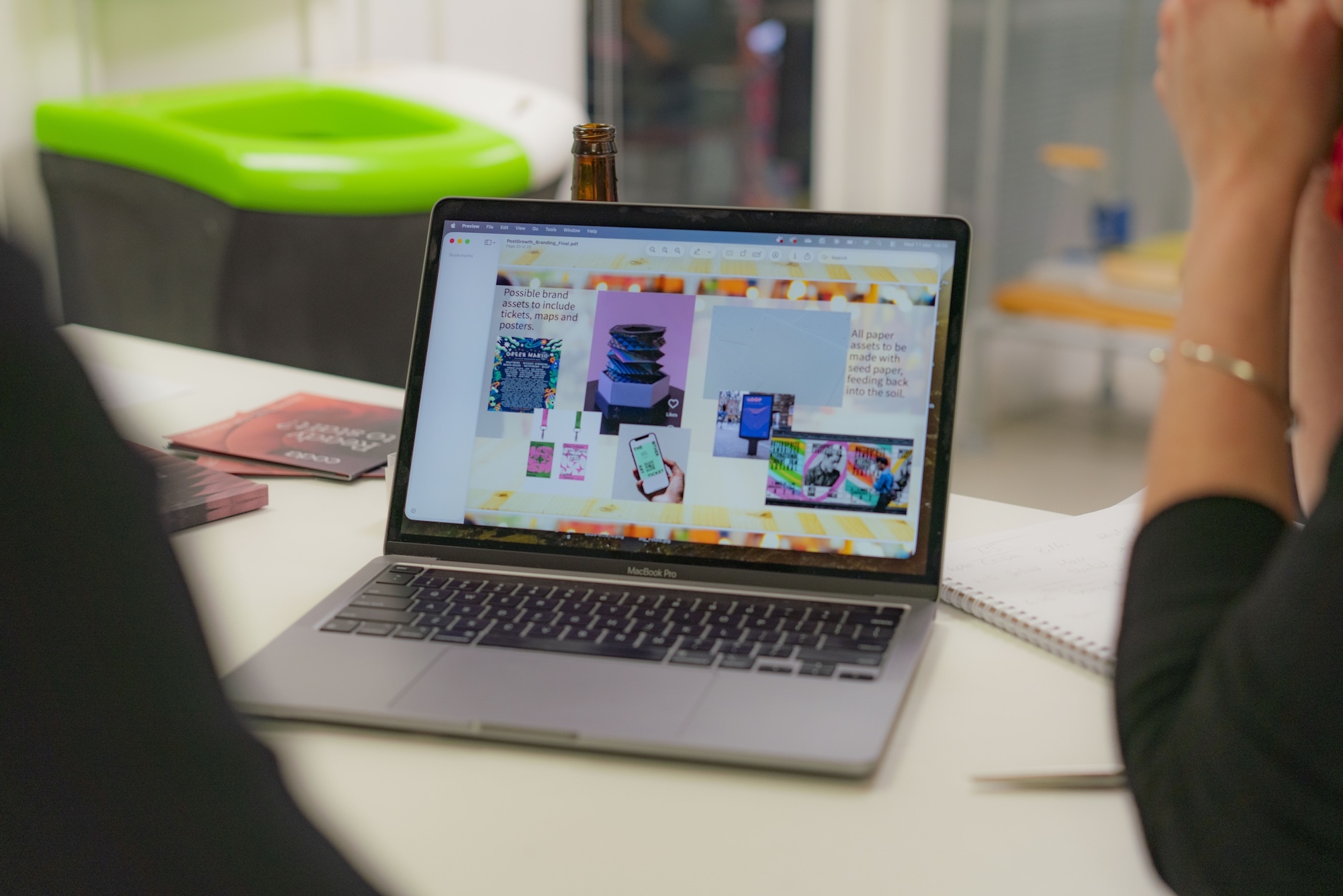 Open MacBook Pro on white desk showing colourful interface with multiple windows. Person's hands visible, green inflatable object in background.