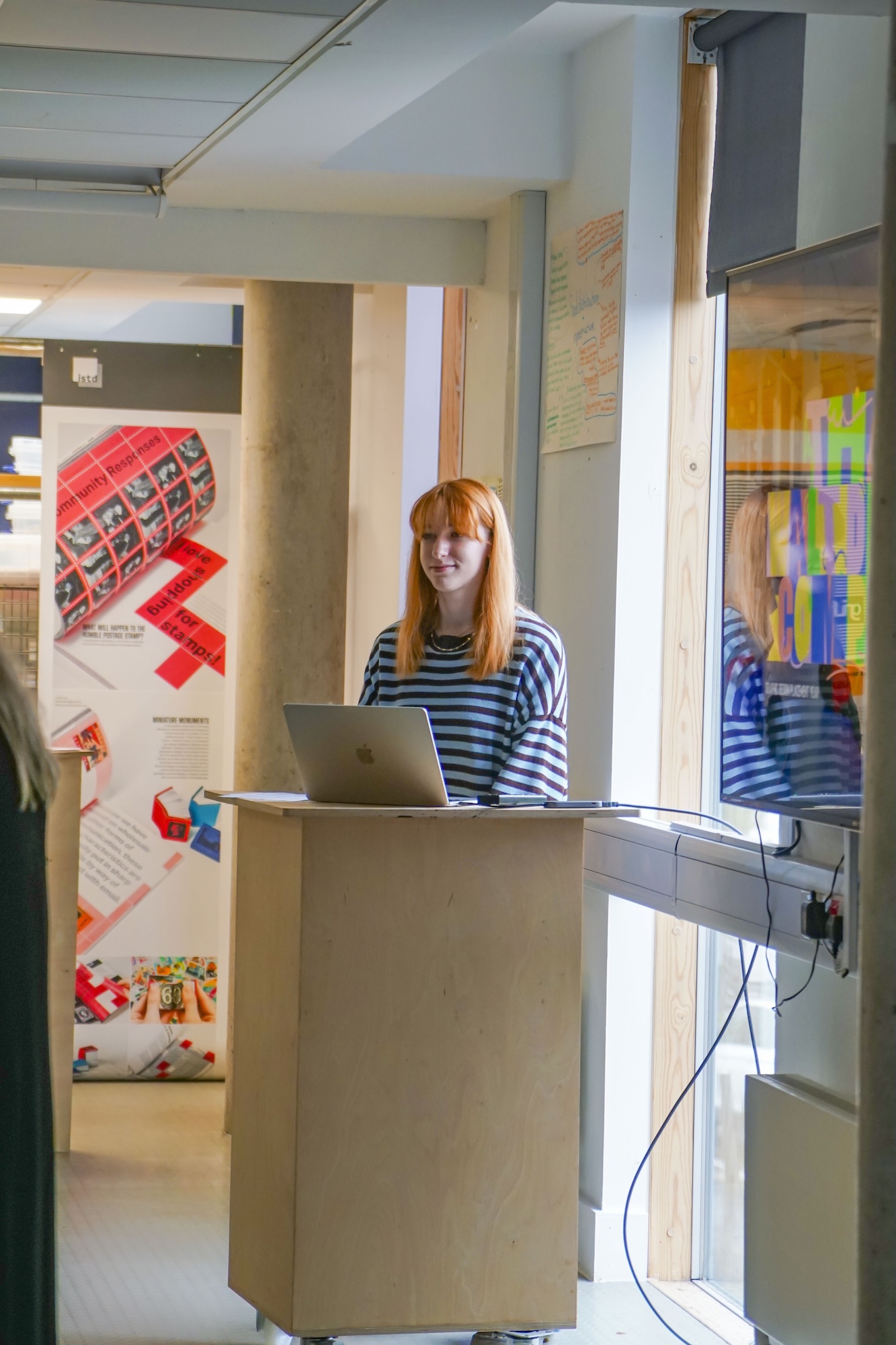 E Jarvis stands behind a laptop on a wooden podium in a bright room, with posters on the wall and a window beside her.