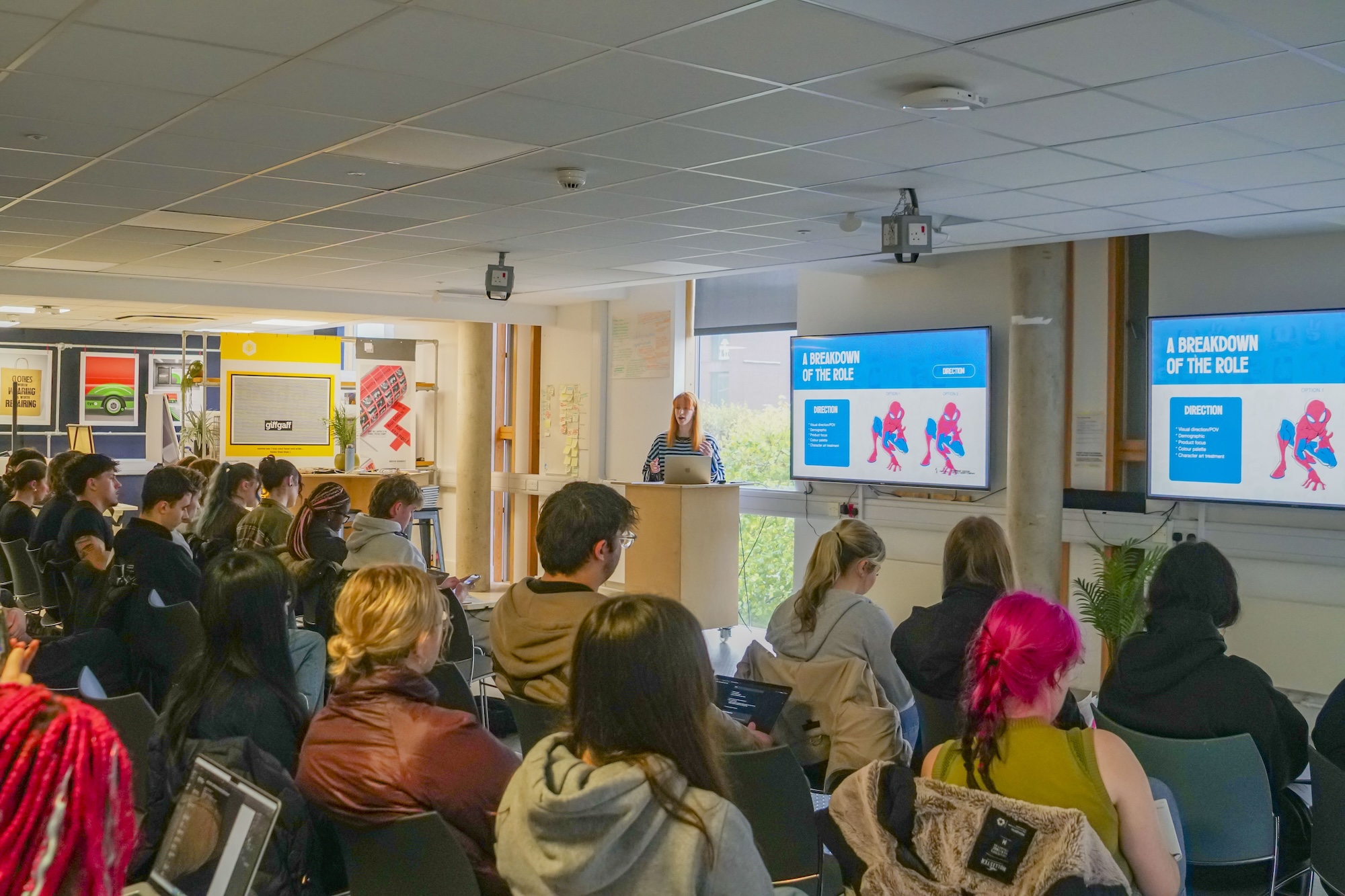 An audience seated in rows at a presentation in a lecture room, with a speaker at a podium beside two large screens showing a slide.