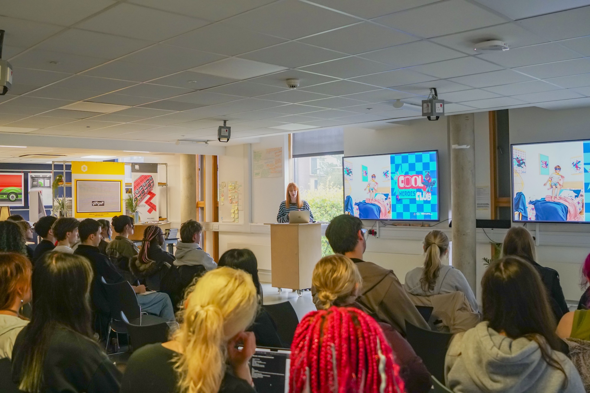 A speaker stands at a lectern in front of an audience seated in rows, with two large presentation screens and a bright windowed wall behind her.