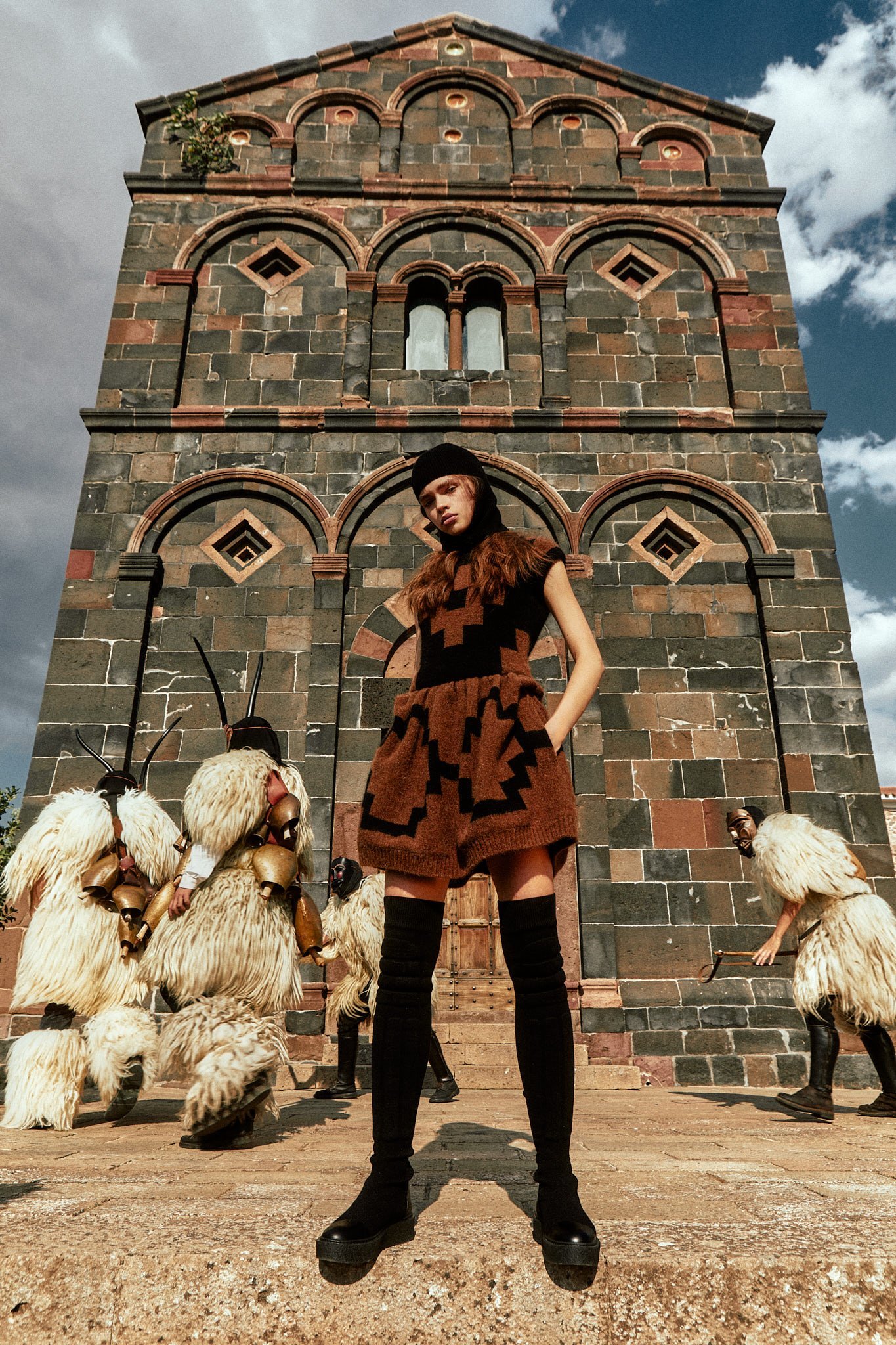 A person posing in front of a church-like structure wearing brown clothing, with people dressed like sheep behind them.