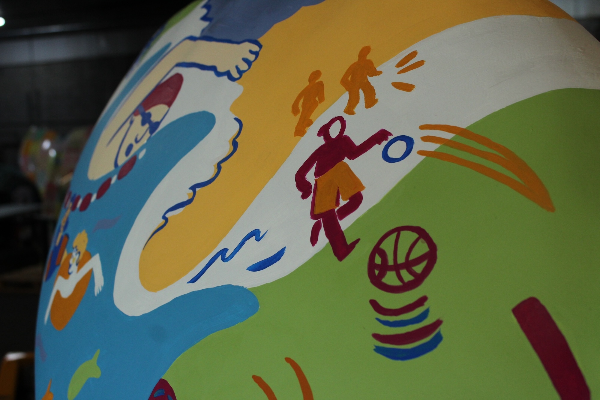 Close-up of colourful illustrations on a mermaid tail sculpture. Illustrations include a person swimming, walking on the beach and playing basketball.