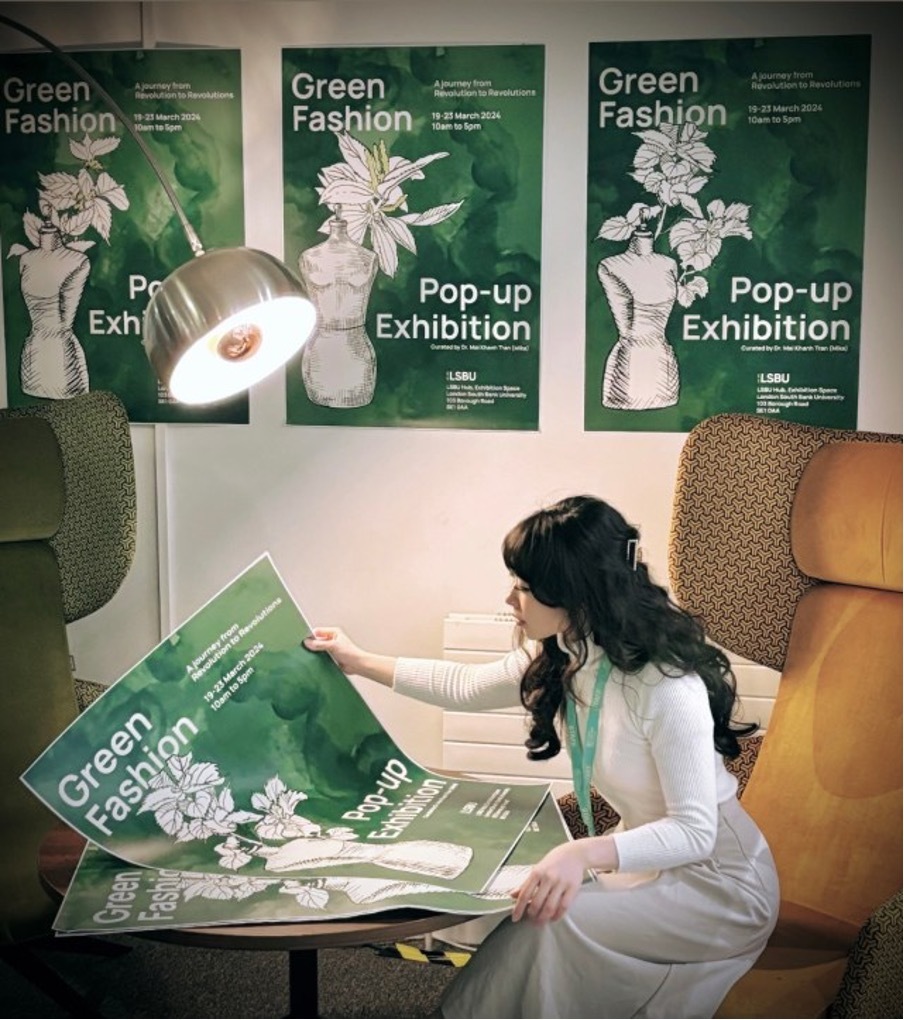 Dr Mika sitting and examining a poster for the Green Fashion pop-up exhibition. Three other copies of the poster are displayed on the wall in the background.
