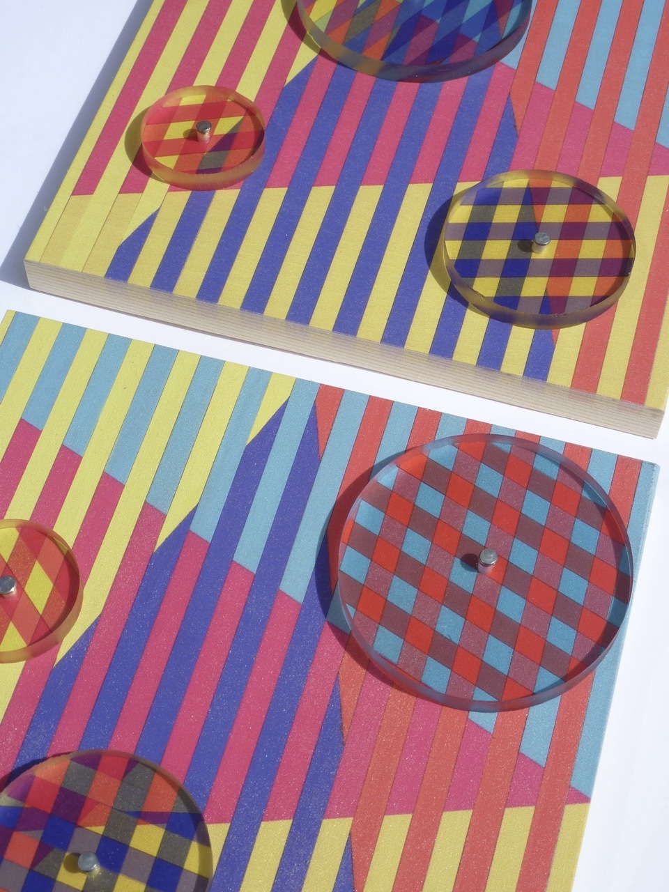 Colourful striped papers with circular transparent overlays creating optical effects. Bright pink, blue, yellow stripes intersect with clear discs.
