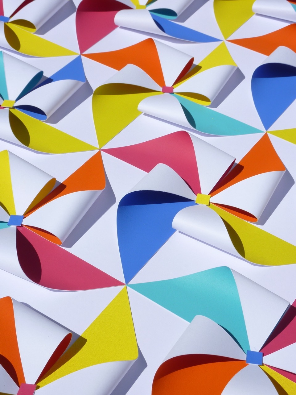 Abstract geometric pattern with overlapping curved triangular shapes in bright colours including yellow, blue, pink, orange and turquoise on white background.